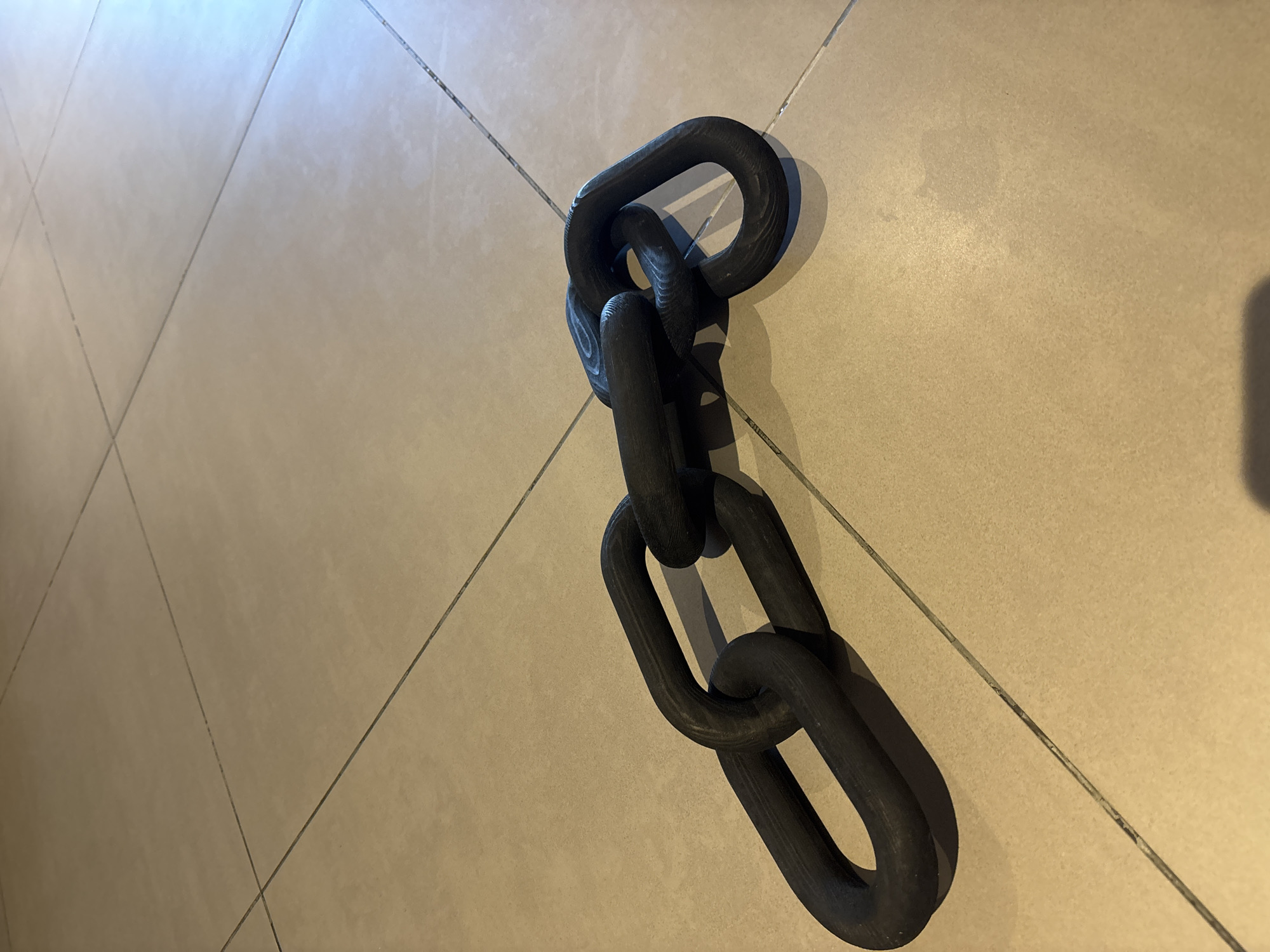 The image shows a close-up, eye-level shot of a heavy-duty black chain lying on a beige tiled floor. The chain consists of several large, interlocking links with a textured surface that appears to be made of wood or a similar material. The lighting suggests an indoor environment, casting shadows on the floor. The tiles are laid in a grid pattern, with visible grout lines. There are no people or other significant objects in the frame. The scene is static and devoid of action. It is not possible to determine the specific location within Dubai, United Arab Emirates, from the visual information provided.