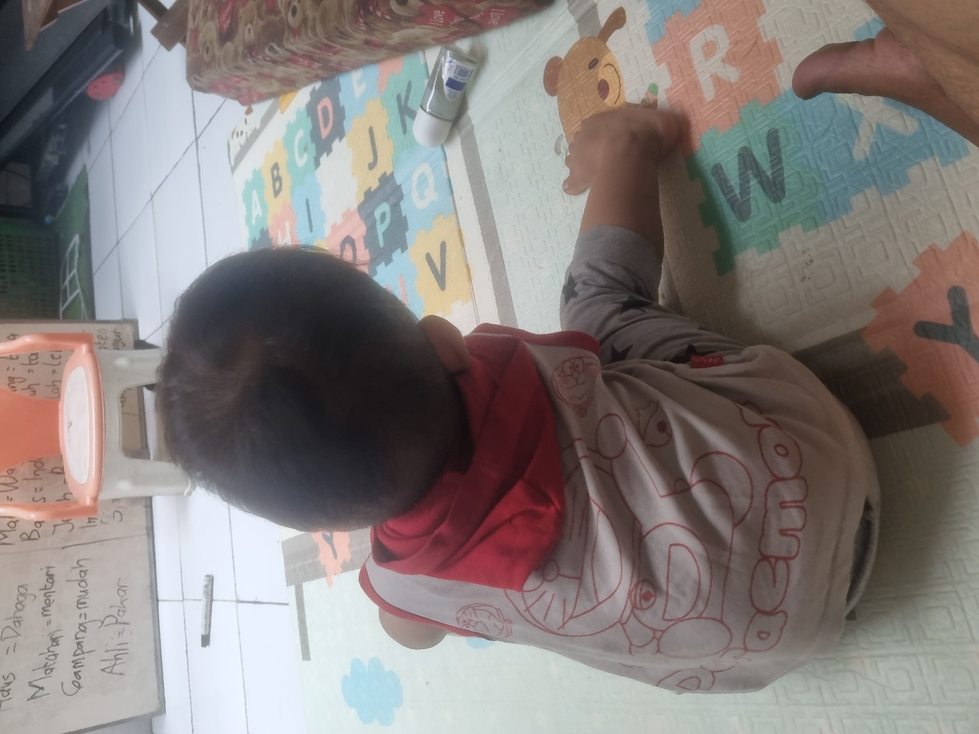 The image shows a young child sitting on a mat with alphabet blocks in what appears to be a domestic setting in Probolinggo, Indonesia. The child is wearing a light-colored t-shirt with a Doraemon design and a red hood. The child's back is to the camera. The mat is a colorful puzzle mat with letters and cartoon bear images. In the background, there is a white board with Indonesian writing, a children's chair, and a treadmill. There is also a small white bottle on the mat. The scene suggests a casual indoor environment, likely a home or play area, in Probolinggo, Indonesia.