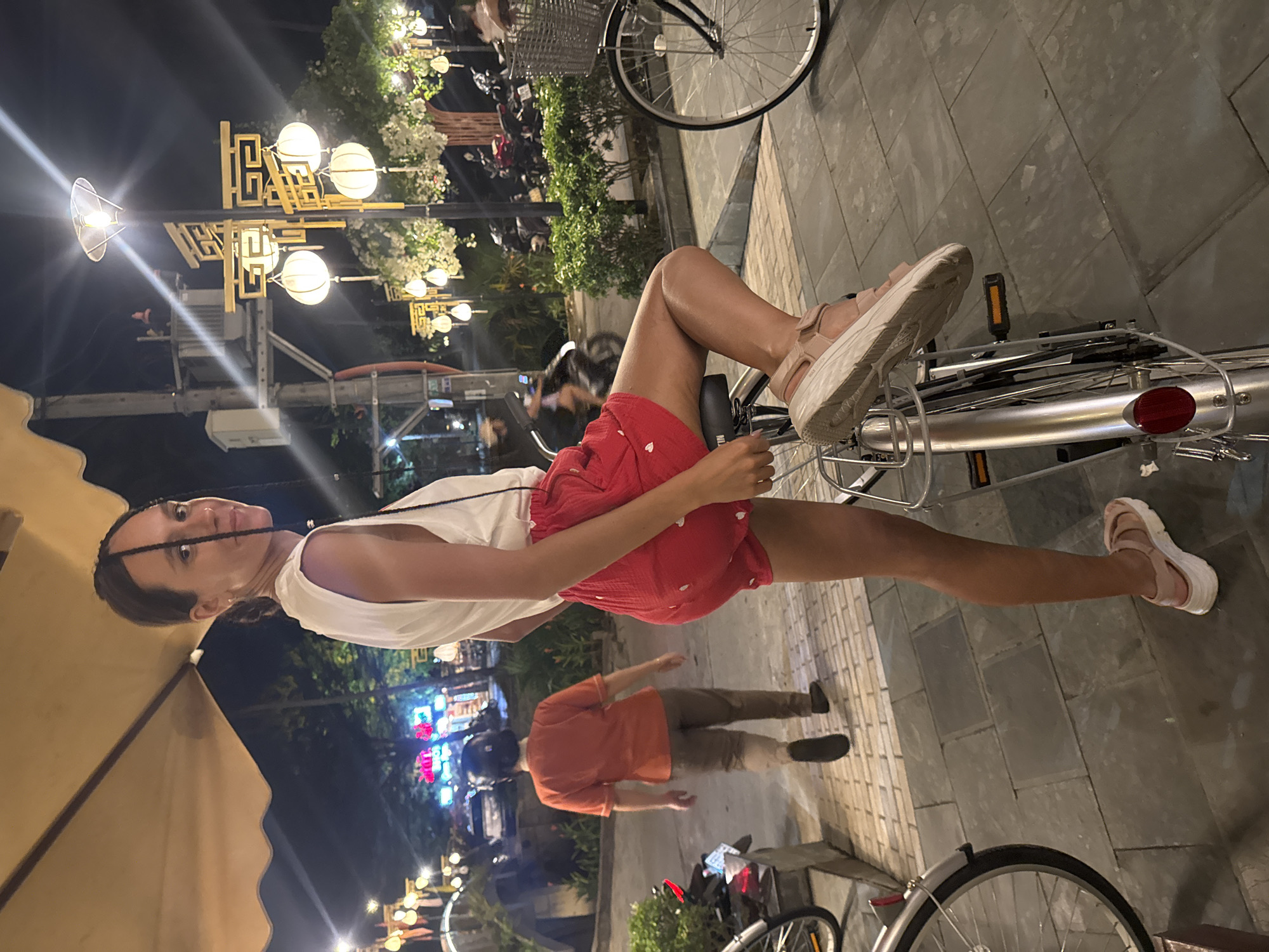 A woman is seated sideways on a bicycle on a paved street in Hội An Ward, Vietnam. She wears a white top, red shorts, and platform sandals. A man in an orange shirt and khaki pants walks behind the bicycle. Streetlights and trees line the street. Other bicycles are visible, and the background shows various buildings with colorful lights.