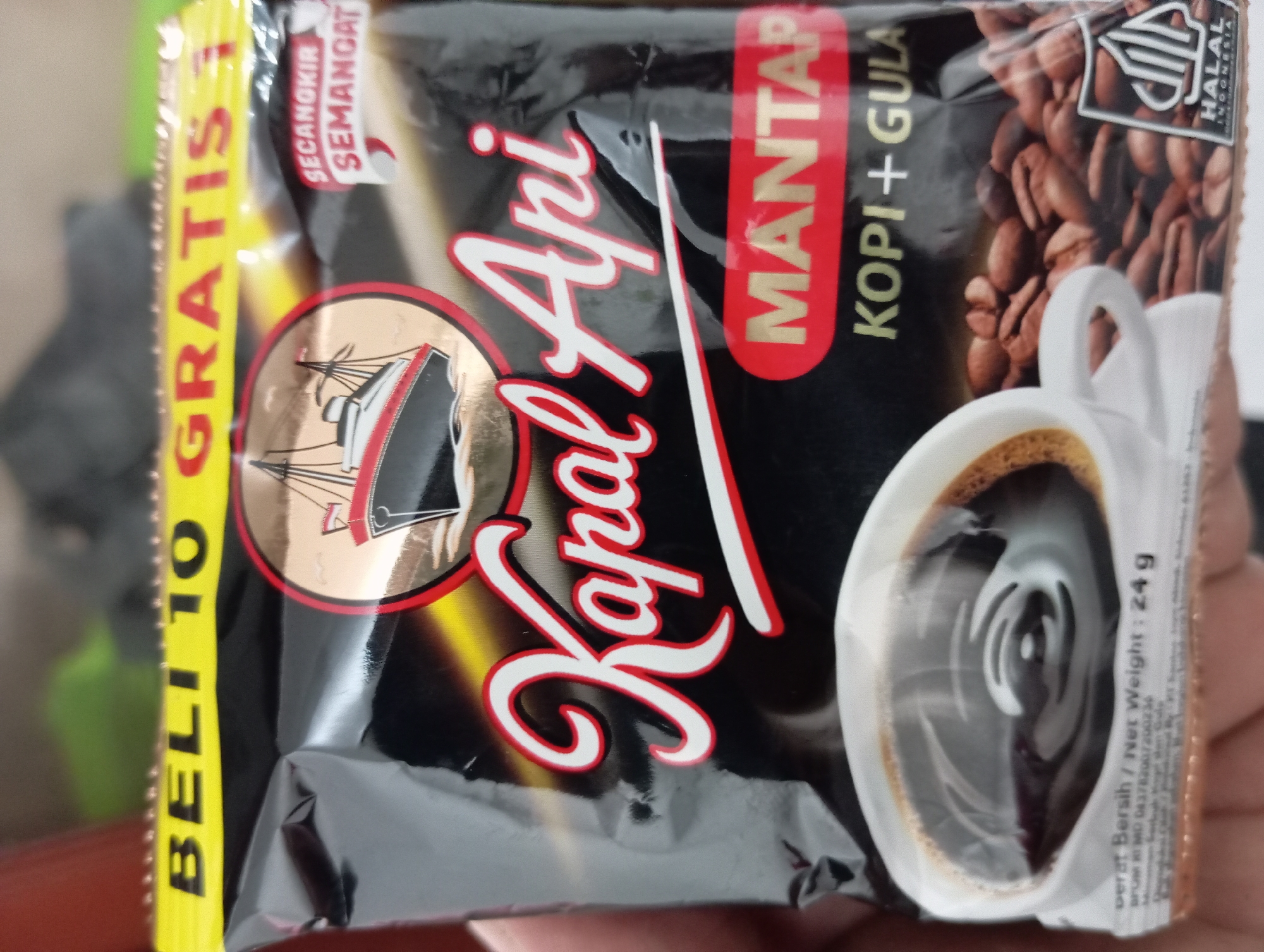 A person's hand is holding a black packet of "Kapal Api" instant coffee, which is a popular Indonesian brand. The packet features a stylized ship logo and text indicating "Mantap Kopi + Gula," which translates to "Great Coffee + Sugar." There's an image of a cup of coffee and coffee beans on the packet, along with a "Halal Indonesia" certification logo. The background suggests an indoor setting with some out-of-focus elements, possibly a table or counter. The image does not provide information about a specific location in Kedungwuluh, Indonesia.