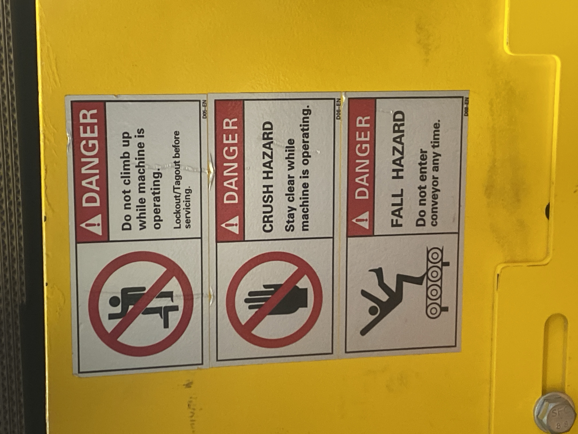 The image presents a close-up view of three industrial safety warning labels attached to a bright yellow piece of machinery or equipment, likely in an industrial or warehouse environment. The image itself is rotated counter-clockwise.
Each label shares a similar design: a prominent red "DANGER" header with an exclamation mark, followed by specific hazard warnings and a graphical pictogram within a red circle with a diagonal line, indicating prohibition. Small alphanumeric codes ("D05-EN," "D06-EN," "D0B-EN") are visible on the top right of each label.
The first label warns of a climbing hazard, stating: "Do not climb up while machine is operating. Lockout/Tagout before servicing." Its pictogram shows a person climbing a ladder with a red prohibitory circle over it.
The middle label indicates a "CRUSH HAZARD" and advises: "Stay clear while machine is operating." The accompanying graphic shows an open hand within a red prohibitory circle.
The third label warns of a "FALL HAZARD" and instructs: "Do not enter conveyor any time." The pictogram depicts a stylized figure falling onto a conveyor belt system.
The yellow surface of the machine shows some wear, scuff marks, and minor dirt. A silver-colored bolt with the marking "SFC 8.8" is visible on the lower right corner of the yellow equipment. No specific time of day, weather conditions, or emotional states are discernible. The image does not provide any visual cues to confirm a location in Paipe, Nigeria, depicting a generic industrial safety scene.