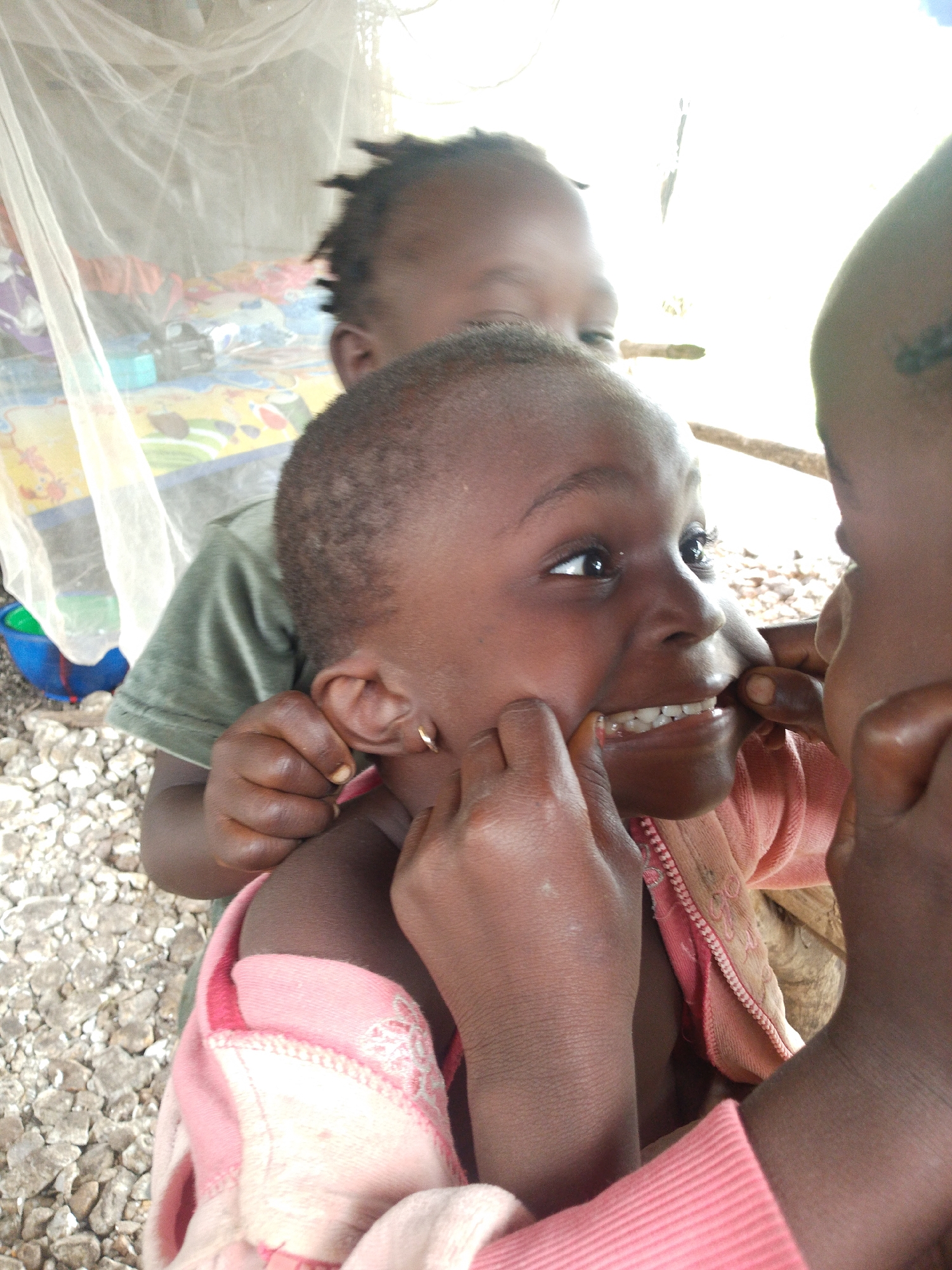 Three young children, likely of African descent, are captured in a close-up shot. The child in the foreground, facing towards the right, has their mouth open, revealing a set of small, white teeth. Another child's fingers are gently pulling on the cheeks of this child, exposing their teeth further. A third child's head is visible in the upper left of the frame, their face obscured. The children are wearing simple clothing: a pink garment with some embroidered detailing is prominent on the child in the foreground, and a green t-shirt is visible on another. The background is slightly out of focus, showing what appears to be a thatched roof structure and a mosquito net. The ground is covered in small, grey stones. The lighting suggests it is daytime, possibly outdoors in a rural or semi-rural setting. The expressions on the visible faces suggest a moment of playful interaction or examination. There is no visible text in the image.