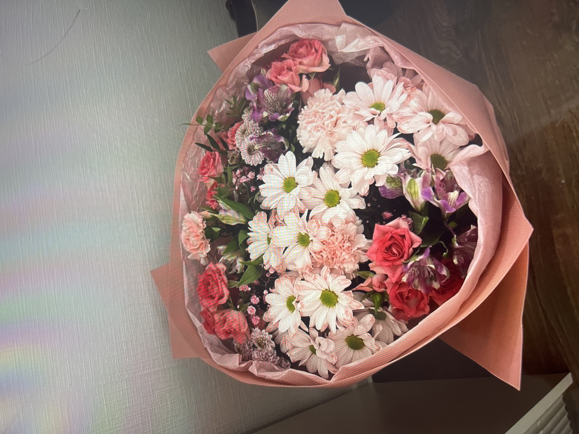 The image shows a bouquet of flowers wrapped in pink paper. The flowers include white daisies, pink roses, and carnations, as well as some purple flowers and green foliage. The bouquet is positioned against a white wall on the left and dark brown surface on the right.