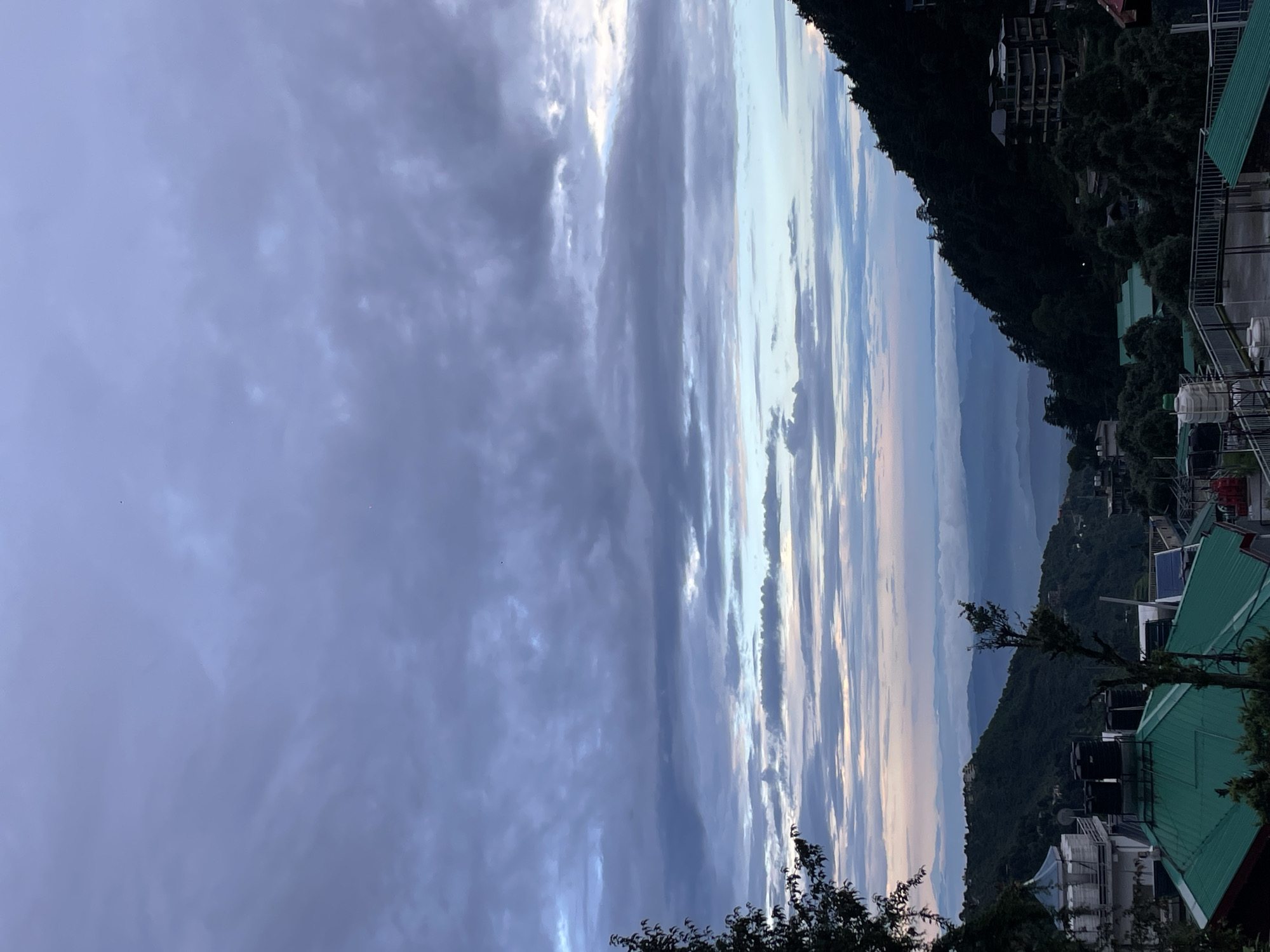 The image captures a panoramic view of a mountainous landscape with a town nestled within the hills, likely in Bhagsu, India, under a cloudy sky. In the foreground, several buildings with green-tiled roofs are visible, some appearing residential or commercial. Water tanks are present on the rooftops of some structures. A few trees, including a prominent one on the left side of the frame, add to the natural elements.
The midground showcases a sloping hillside densely covered with dark green trees, typical of a mountain region. Scattered amidst the foliage are more buildings, some appearing to be multi-story structures, suggesting a developed settlement on the slopes. Further in the distance, layers of mist or low-lying clouds create a soft, hazy effect, obscuring the base of more distant mountains.
The background is dominated by a vast expanse of sky filled with a variety of clouds. The clouds are layered, with darker, more turbulent formations towards the top, transitioning into lighter, more wispy patterns closer to the horizon. Faint traces of pink and blue hues suggest either early morning or late evening light filtering through the clouds. The overall atmosphere is serene and a bit melancholic due to the overcast sky and diffused light. No people are clearly identifiable in the scene.
