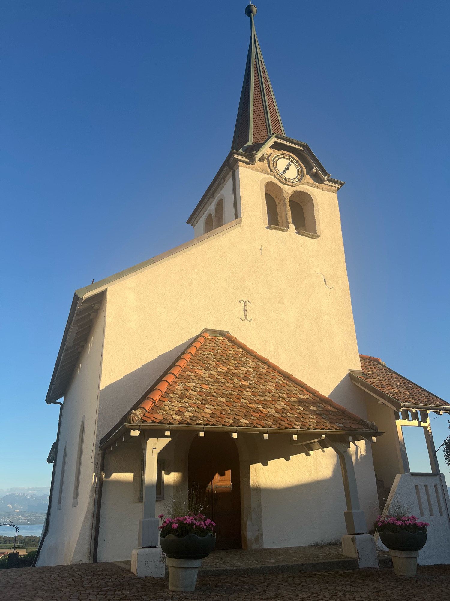 The image shows the Église Saint-Jean-Baptiste, a church located in Féchy, Switzerland. The church is built of light-colored stone and features a tall steeple with a clock. A red-tiled roof extends over the entrance, supported by white pillars. Flower pots with pink blossoms are placed on either side of the entrance. The church is bathed in the warm light of the setting sun, casting long shadows on the ground. In the background, a body of water and distant mountains can be seen, suggesting a scenic location overlooking Lake Geneva. The sky is a clear, bright blue. There are no people visible in the image.