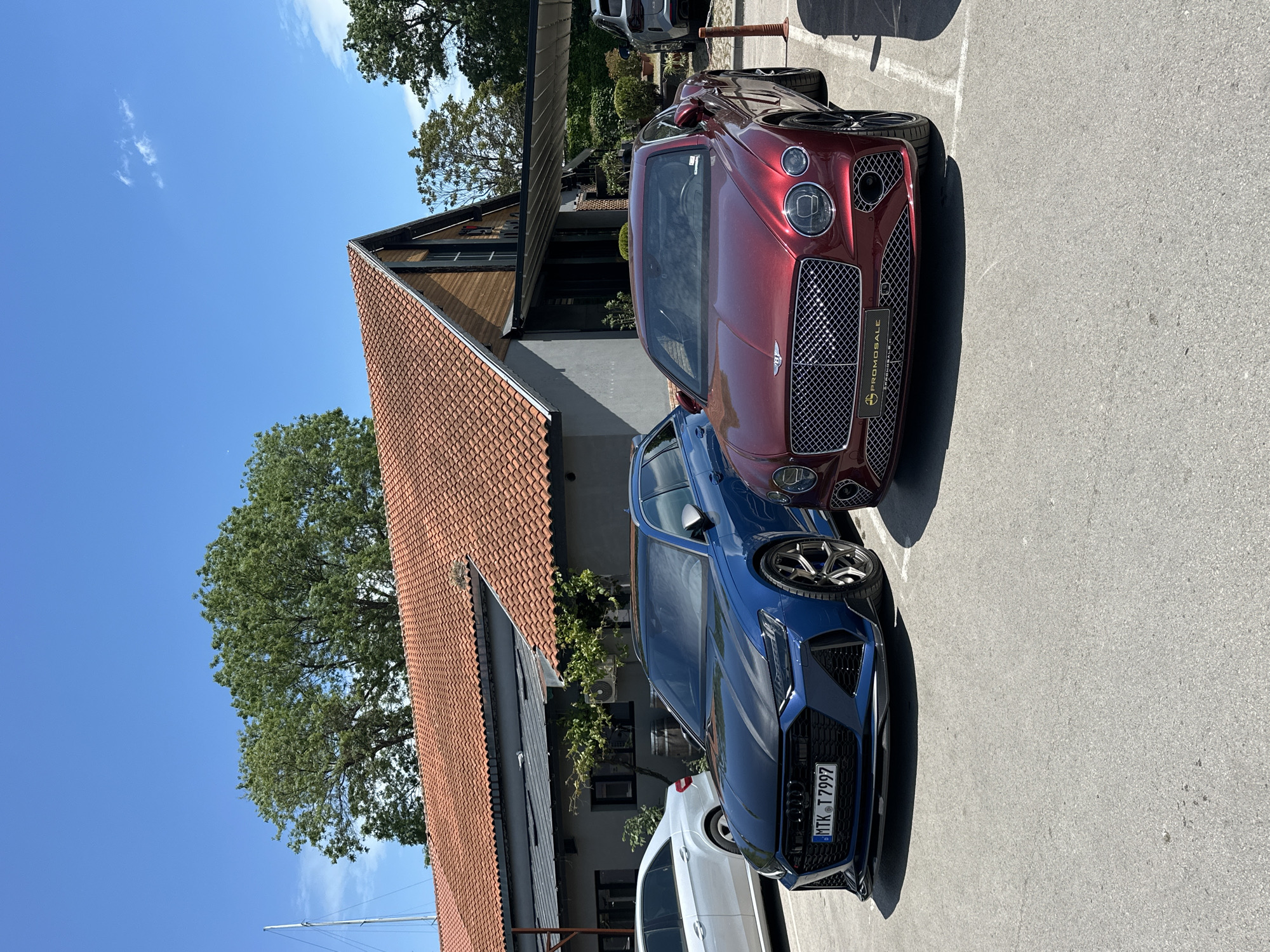 The image shows a parking lot in Varna, Bulgaria, with a red Bentley and a blue Audi RS6 parked next to each other. Behind the cars is a building with a red tile roof and a light-colored facade. A tree is visible to the left of the building. The sky is blue with a few clouds.