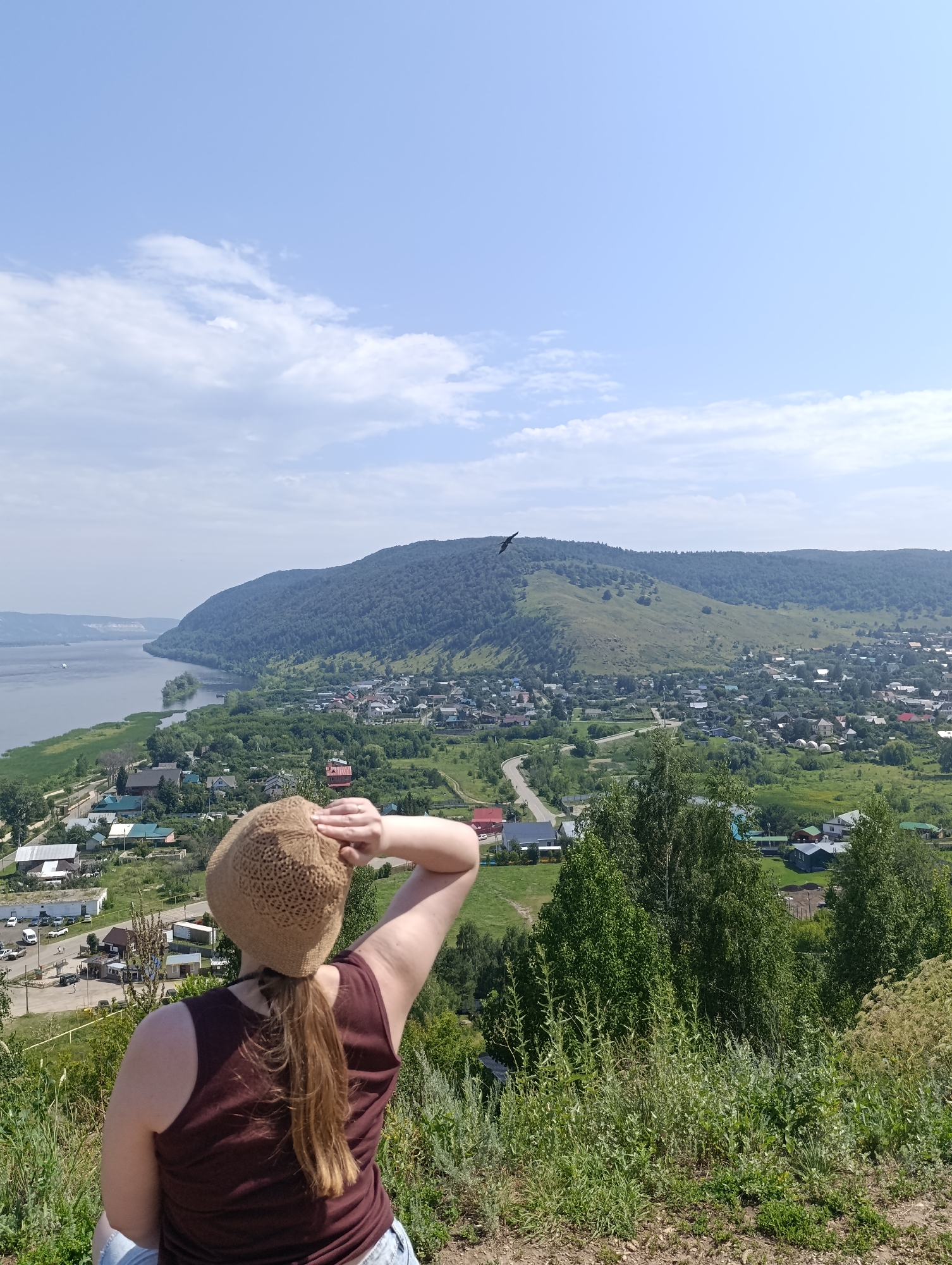 A person wearing a brown, crocheted hat and a maroon tank top is standing on a hillside, looking out over a valley. The person's right arm is raised, with their hand shielding their eyes from the sun. The valley below contains a river, a town with numerous buildings, and rolling, tree-covered hills in the distance. A bird is visible flying in the sky above the town. The sky is blue with scattered white clouds. The foreground is dominated by green vegetation and dry, rocky ground. The scene appears to be the city of Solnechnaya Polyana, Russia, likely taken from a scenic overlook.