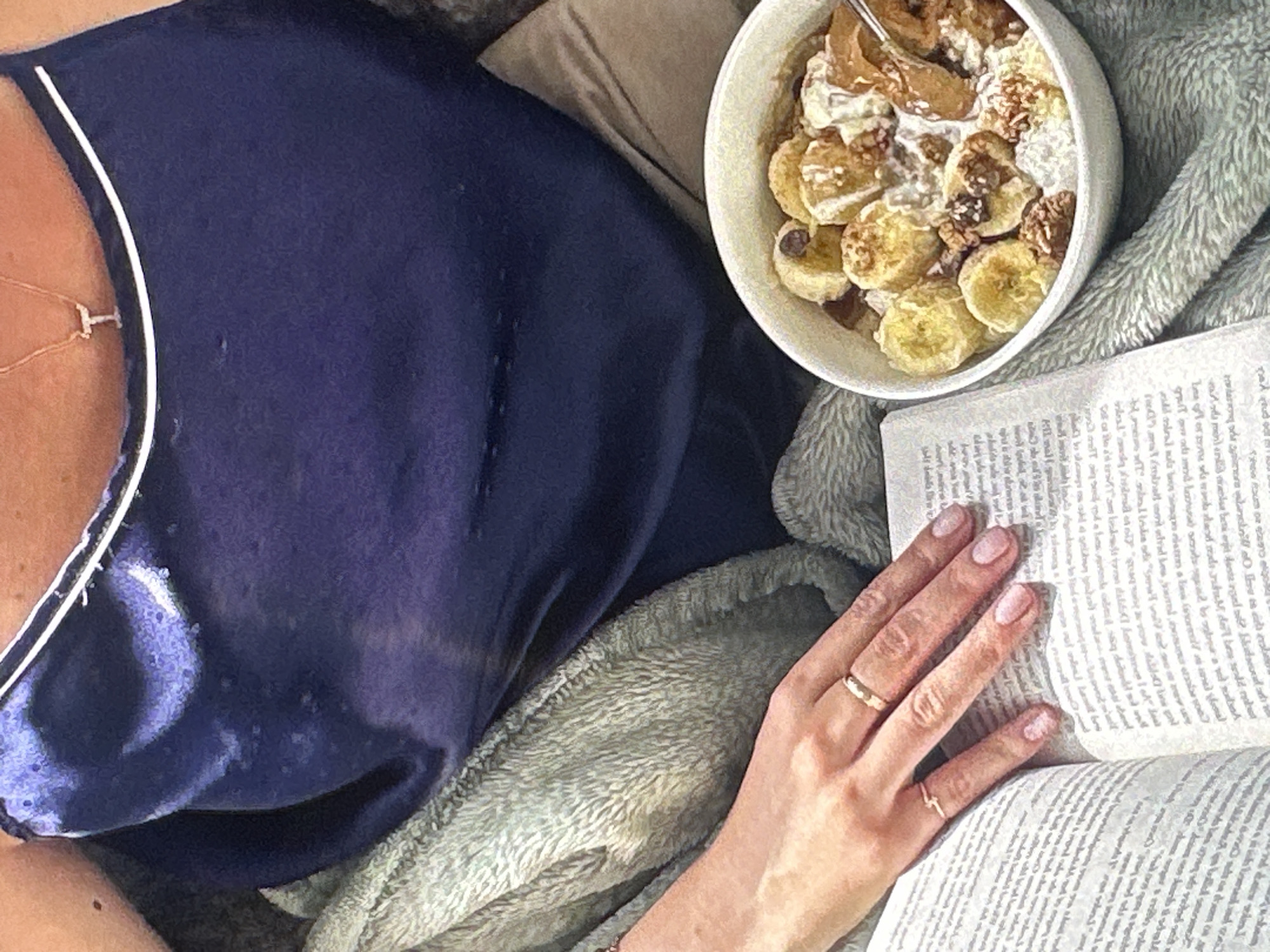 A person wearing a navy blue top with a white trim is lying down with a bowl of sliced bananas, nuts, and a light-colored liquid. The person is holding a book open with their right hand, which has two rings on its fingers. The person is covered by a light green blanket. The scene is indoors. There is no evidence to suggest the location is in Yerevan, Armenia.