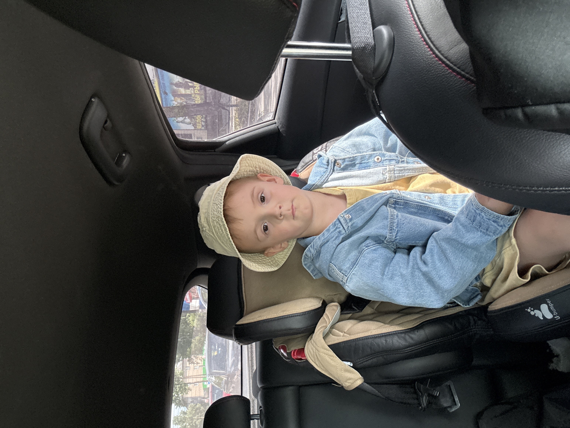 A young boy wearing a bucket hat, a denim jacket, and a yellow shirt sits in a car seat in the back of a vehicle. He is facing forward and looking at the camera. Through the rear window, a street scene is visible, including buildings, trees, and other cars. The image appears to have been taken in the city of Chaikovsky, Russia. The boy is positioned in the center of the frame, with the car's interior filling the foreground and background. The lighting suggests it is daytime, and the overall mood is casual and candid.