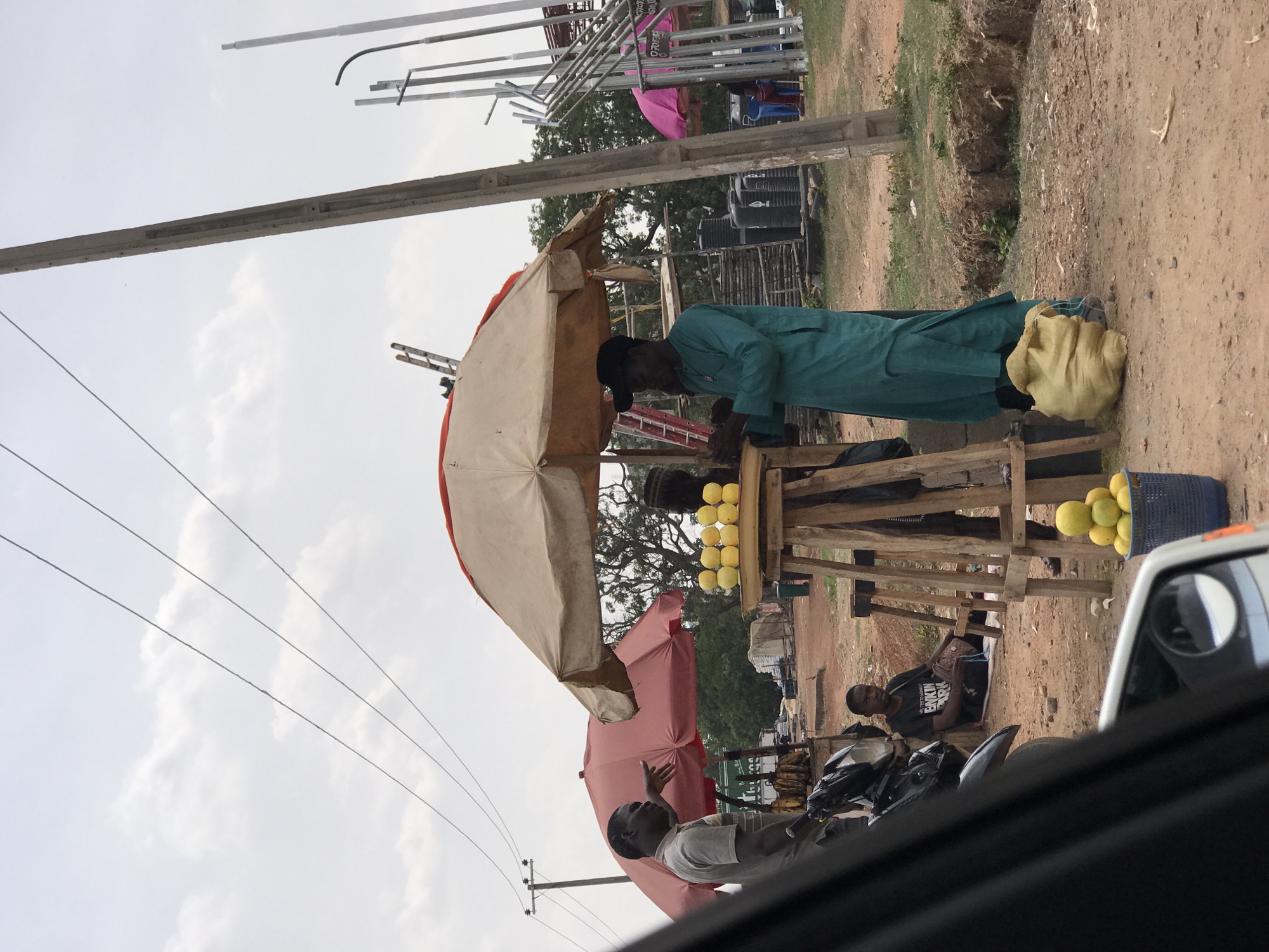 An outdoor scene in Abuja, Nigeria, captures a bustling roadside market area under a partly cloudy sky. The foreground is dominated by unpaved, dusty ground.
Three men are visible. In the center, a man wearing a teal-green tunic and a dark cap stands behind a wooden stall, seemingly tending to his goods. Next to him on the ground is a large, yellow sack. To his left, another man in a white t-shirt stands and gestures with his hand, possibly engaged in conversation or drawing attention. Further to the right and slightly behind, a third man sits on the ground near a black motorcycle. He wears a dark t-shirt with the text "I'M TRYING TO ORGANIZE MY LIFE. BIKIN' ARK" printed on it.
Market stalls made of wooden frames are set up along the roadside. Several baskets and crates filled with bright yellow fruits, possibly oranges or grapefruits, are displayed on and around these stalls. Two large parasols provide shade: one beige, covering the main stall, and another pink/red one further down the road.
In the background, there are trees, some black water tanks, and metal scaffolding structures. A concrete utility pole and power lines stretch across the upper left portion of the image. The photo appears to be taken from inside a vehicle, with part of a white car's door frame and side mirror visible in the bottom right corner. The scene depicts a typical daytime market activity, with a generally neutral mood.
