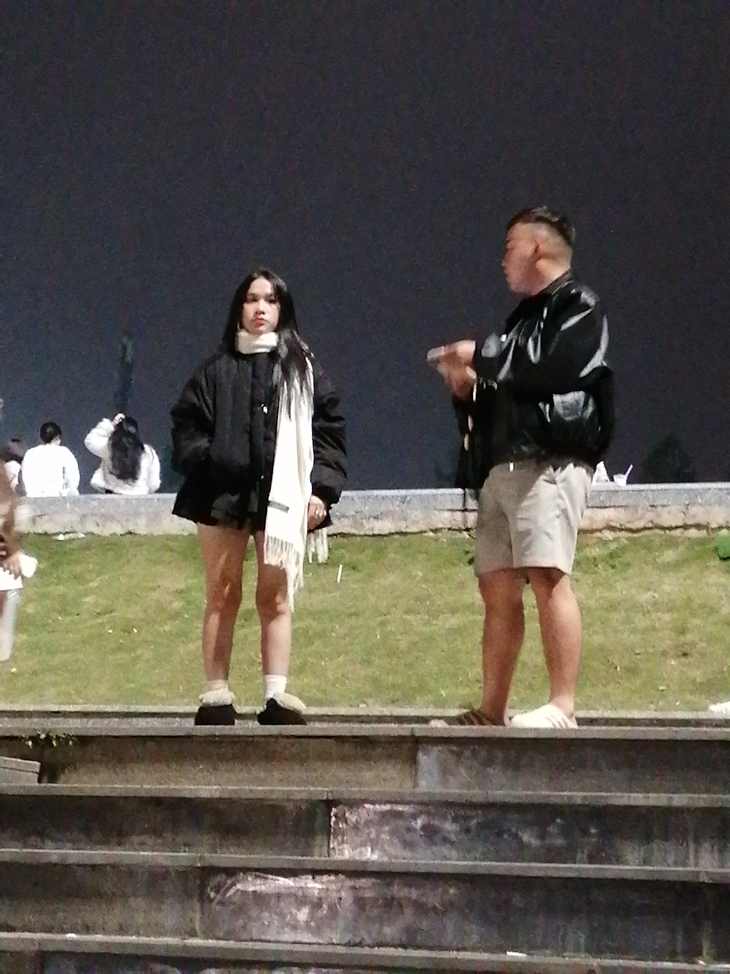The image depicts two individuals standing on a grassy embankment at night, with a dark background suggesting an outdoor urban setting, possibly Da Lat, Vietnam, given the presence of stairs and manicured landscaping. On the left is a young woman wearing a black puffer jacket, a white scarf, and dark shorts. She has long, dark hair and is looking towards the viewer. On the right is a young man in a black leather jacket, grey shorts, and white slide sandals. He is facing the woman and appears to be holding a mobile phone or a similar object in his hand, gesturing towards her. The ground in front of them is a grassy area, and behind them, a low wall or railing is visible, with other people in the distance. The foreground shows a set of concrete stairs. The lighting suggests artificial illumination, likely from streetlights or nearby buildings.