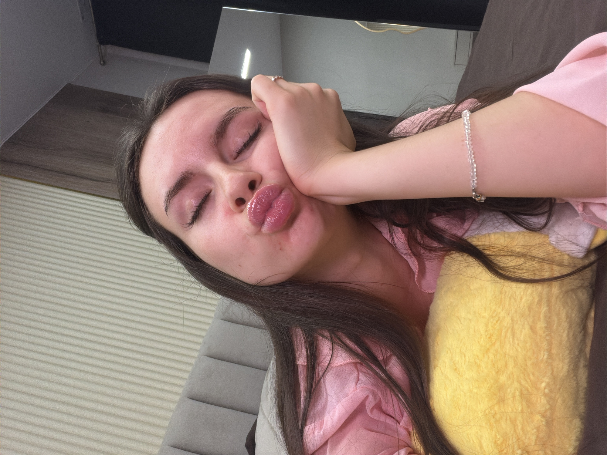 A young adult female with long, dark hair is depicted in a close-up, horizontal orientation, lying on a bed. Her eyes are closed, and her lips are pursed forward in a glossy, light pink pout. Her right hand supports her head, with her elbow resting on the bed surface. A silver-toned ring is visible on her right ring finger, and a clear, faceted beaded bracelet adorns her right wrist. She wears a light pink, long-sleeved shirt. A soft, plush, bright yellow object, possibly a pillow or stuffed toy, is situated beneath her left arm and chest area.
The background indicates an indoor setting, likely a bedroom. To the upper left, a gray, upholstered headboard with vertical channel tufting is visible, positioned against a light-colored wall featuring subtle vertical fluting or ridging. To the upper right, a section of dark furniture, a dark curtain rod, and a dark curtain are partially visible. A large rectangular mirror reflects an interior space with a gray wall and a linear ceiling light fixture. The bed itself appears to be covered with dark brown or gray bedding.