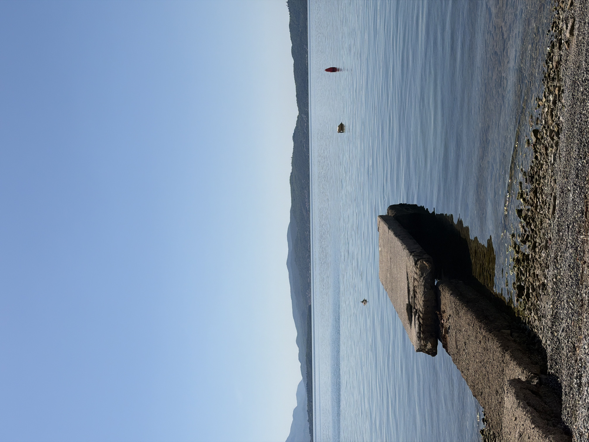 The image depicts a coastal scene in Bijela, Montenegro. The camera is positioned at the edge of the shoreline, looking towards the water. A weathered concrete structure extends into the water from the rocky beach. In the distance, a tree-covered hill appears. In the water, two objects are visible: one red and one yellow. The sky is clear and blue, and the water reflects the sky's color. The overall scene is calm and serene.