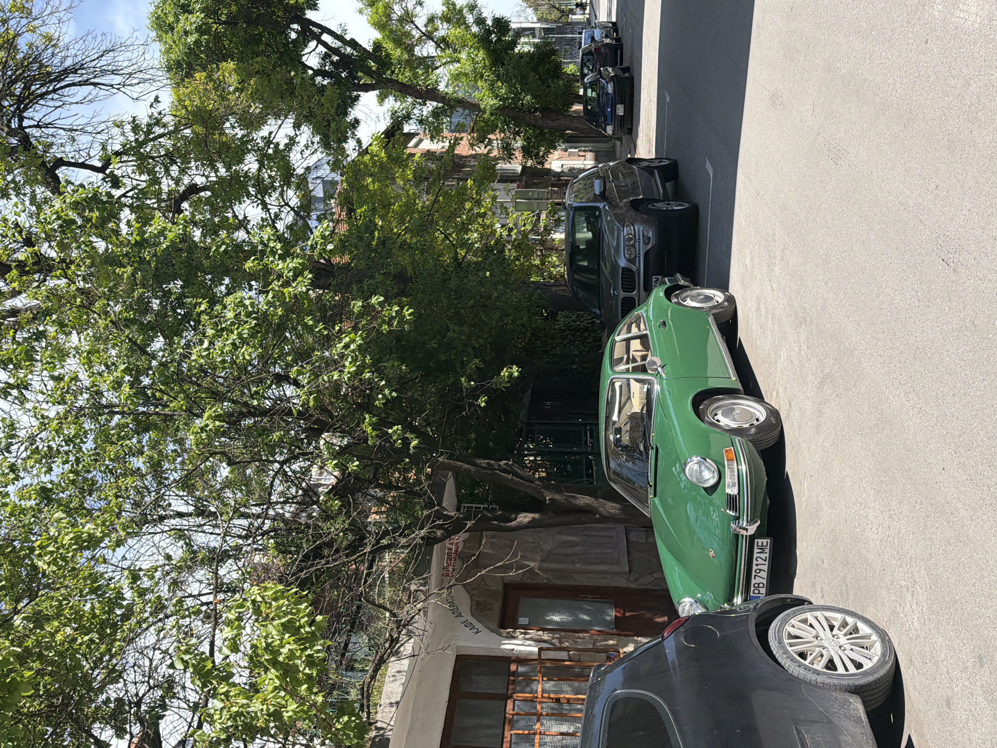 The image depicts a street scene in an unidentified city, possibly during daylight hours given the sun's position. On the right side of the frame, cars are parked along the curb, including a vintage green car with the license plate "PB 7912 ME", a dark-colored BMW, and other vehicles lined up further down the street. On the left side, there is a building partially obscured by a large tree with thick green foliage. An inscription with "KADE ABEPAT" is visible on the building's facade. The environment appears to be residential with a mix of natural elements and urban infrastructure.