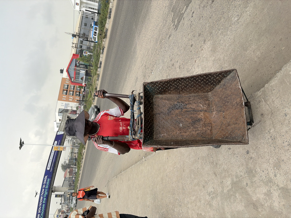 The image presents an urban street scene in Jakande, Nigeria, under a light grey, overcast sky. In the foreground, a dark-skinned individual, appearing male, is positioned behind a large, empty, two-wheeled handcart. He wears a red short-sleeved jersey with white trim (an Arsenal football club shirt with "Emirates Fly Better" sponsor), dark trousers, and a dark, wide-brimmed hat that casts a shadow over his face. His hands grasp the dark metal handles of the cart, which features a rusted, dark brown diamond-plate metal bed.
To the immediate left, a portion of a second individual is visible, wearing a striped tan and white short-sleeved shirt. Further along the concrete sidewalk, a third individual, a dark-skinned female, walks away from the viewer. She wears an orange short-sleeved top, black shorts, and white sneakers, carrying a white plastic bag in her left hand.
The scene includes a multi-lane paved road running parallel to the sidewalk. A pedestrian bridge with an overhead gantry sign spans the road in the mid-ground. The sign displays prominent white text: "NOW YOU KNOW HOW TO TRADE. CHOOSE THE RIGHT BROKER." Various multi-story buildings, some residential-style with orange and white facades, and others commercial with a red roofline, are visible across the road. Vehicles, including a light-colored sedan and a white and blue van, are present on the road in the distance. Streetlights on poles are also visible above the road.
