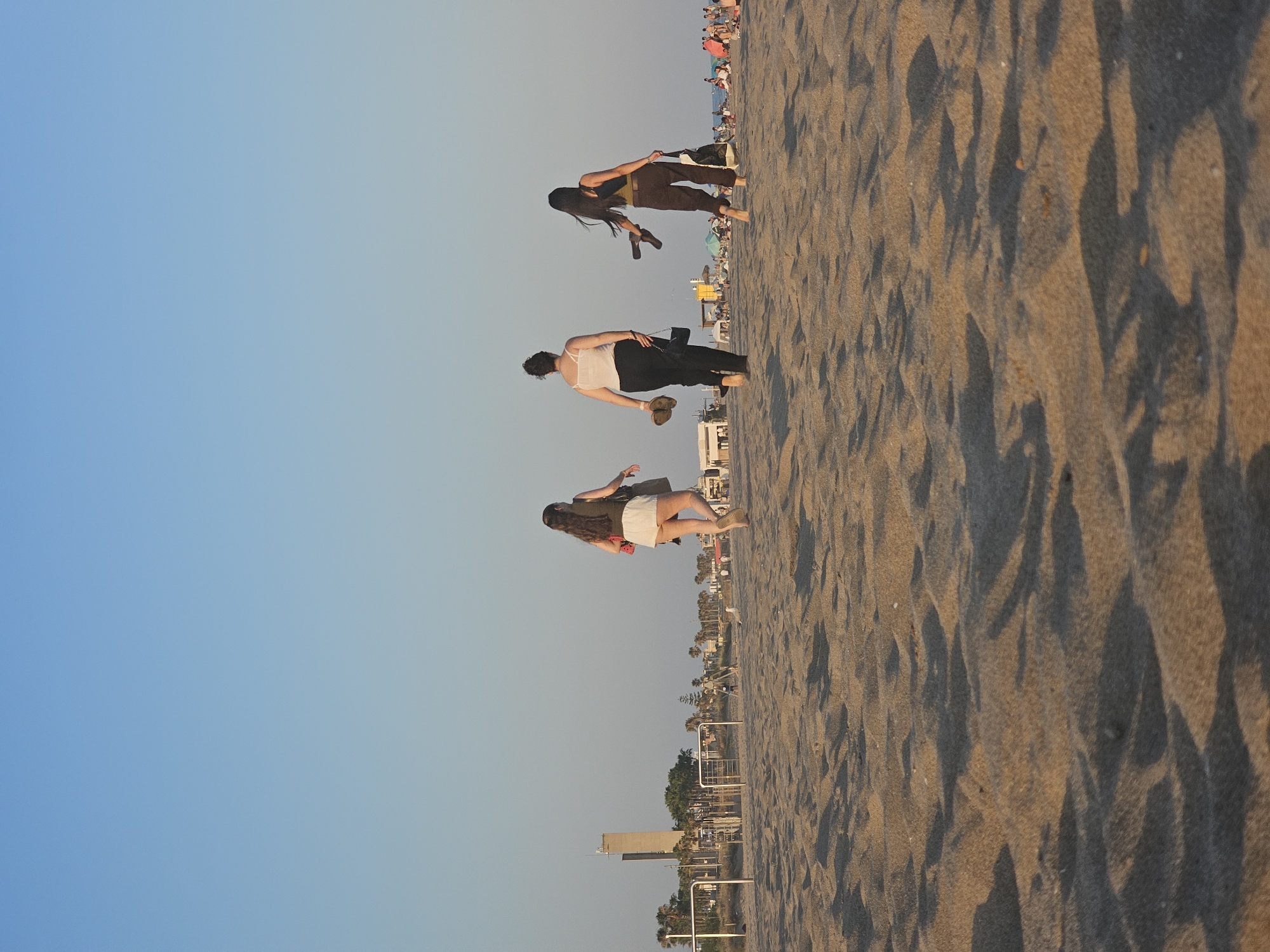 Three women walk barefoot on a sandy beach towards the water at sunset in Castelldefels, Spain. The woman on the left wears a white top and white shorts, and carries a brown purse. The woman in the middle wears a white tank top and black pants, and carries a black purse. The woman on the right wears a dark tank top and dark pants, and carries a light-colored bag and a pair of dark sandals. In the background, a tall, slender tower stands near palm trees and other buildings. The sky is clear and blue, with a gradient of lighter blue towards the horizon. The sand is rippled, with shadows cast by the setting sun.
