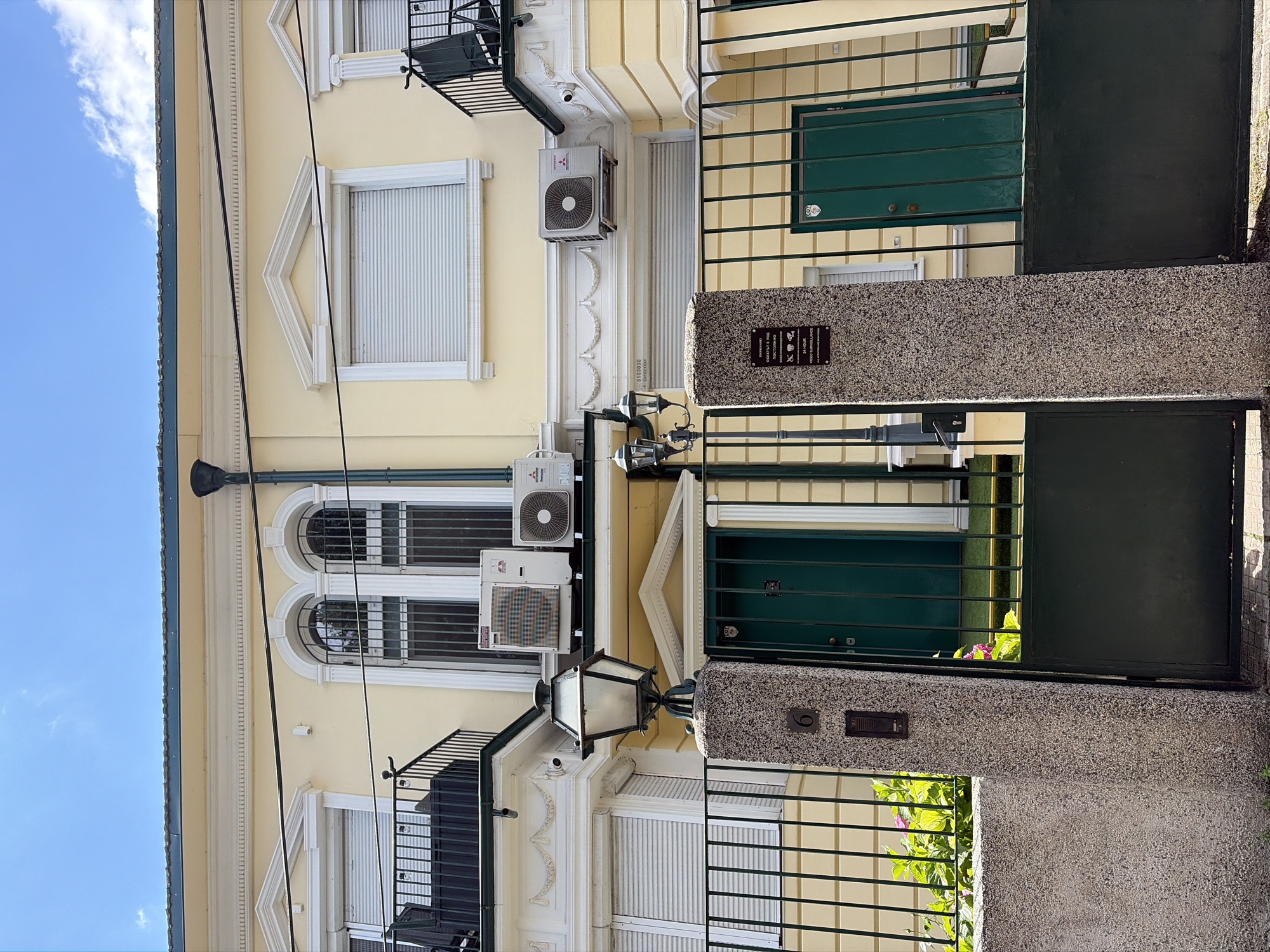 The image depicts the facade of a building in Sofia, Bulgaria. The building is painted in a pale yellow hue with white decorative elements. On the upper level, there are arched windows with metal grates and white roller shutters. Air conditioning units are mounted on the exterior wall.
The ground level features a large, dark green metal gate. To the left of the gate, there is a stone pillar with the number "6" engraved on it and an intercom system. A lamppost with a lantern is positioned on the pillar. A sign on the pillar reads in Russian and English "24 HOUR VIDEO SURVEILLANCE. ALL ACTIVITIES ARE RECORDED."
To the right of the gate, another dark green door is visible, also behind a metal railing. Plants with pink and green foliage are growing near the base of the pillar and gate. The sky above is blue with some scattered clouds. The overall impression is of a residential or office building with security measures in place.