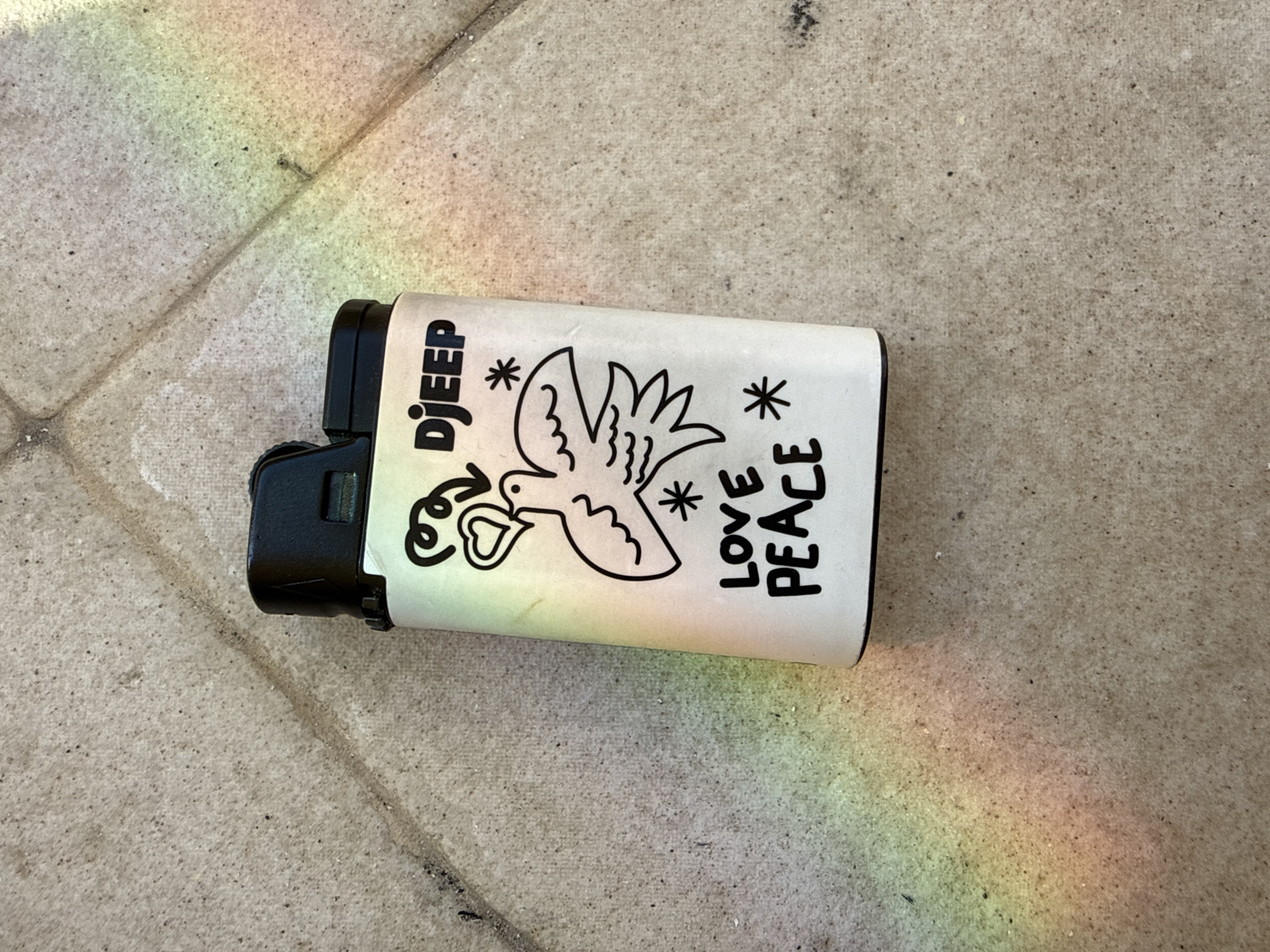 A close-up photograph depicts a Djeep brand lighter resting on a stone-tiled surface. The lighter is white with black text and illustrations. It has a drawing of a dove holding a heart in its beak, along with stars and the words "LOVE PEACE." A rainbow-colored light streak crosses the stone surface. The location is likely a sidewalk or patio in an outdoor setting in Bar, Montenegro, given the architectural style of the tile and the presence of the rainbow.