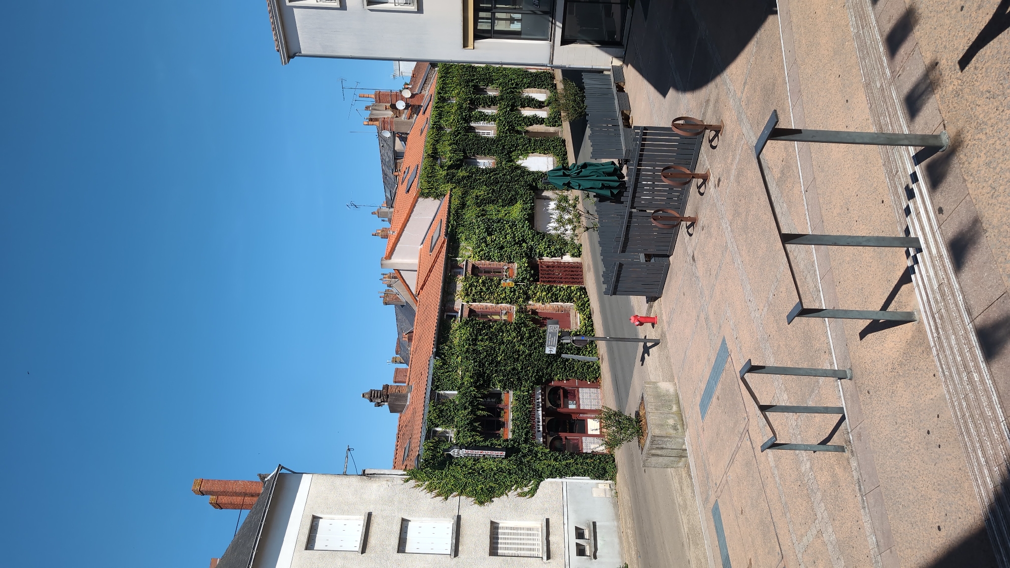 The image depicts a street scene in the city of Cholet, France, likely in a commercial or residential area with older architecture. In the foreground, a set of concrete stairs with metal handrails leads up from a tiled plaza. To the left and right of the plaza are buildings. The building on the left is a light-colored, multi-story structure with white-shuttered windows. The building in the center, which dominates the middle ground, is an older, brick building extensively covered in green ivy. It features multiple windows and an arched doorway at ground level, with signage that appears to indicate a business. A red fire hydrant is visible near the sidewalk in front of this ivy-covered building. Beyond the ivy-covered building, other buildings with red-tiled roofs and chimneys are visible. A dark grey fence with decorative circular metal elements encloses an outdoor seating area in front of one of the buildings, and a green umbrella is set up within this area. The sky is clear and blue, suggesting a sunny day. The overall impression is one of a charming, European street with historical buildings.