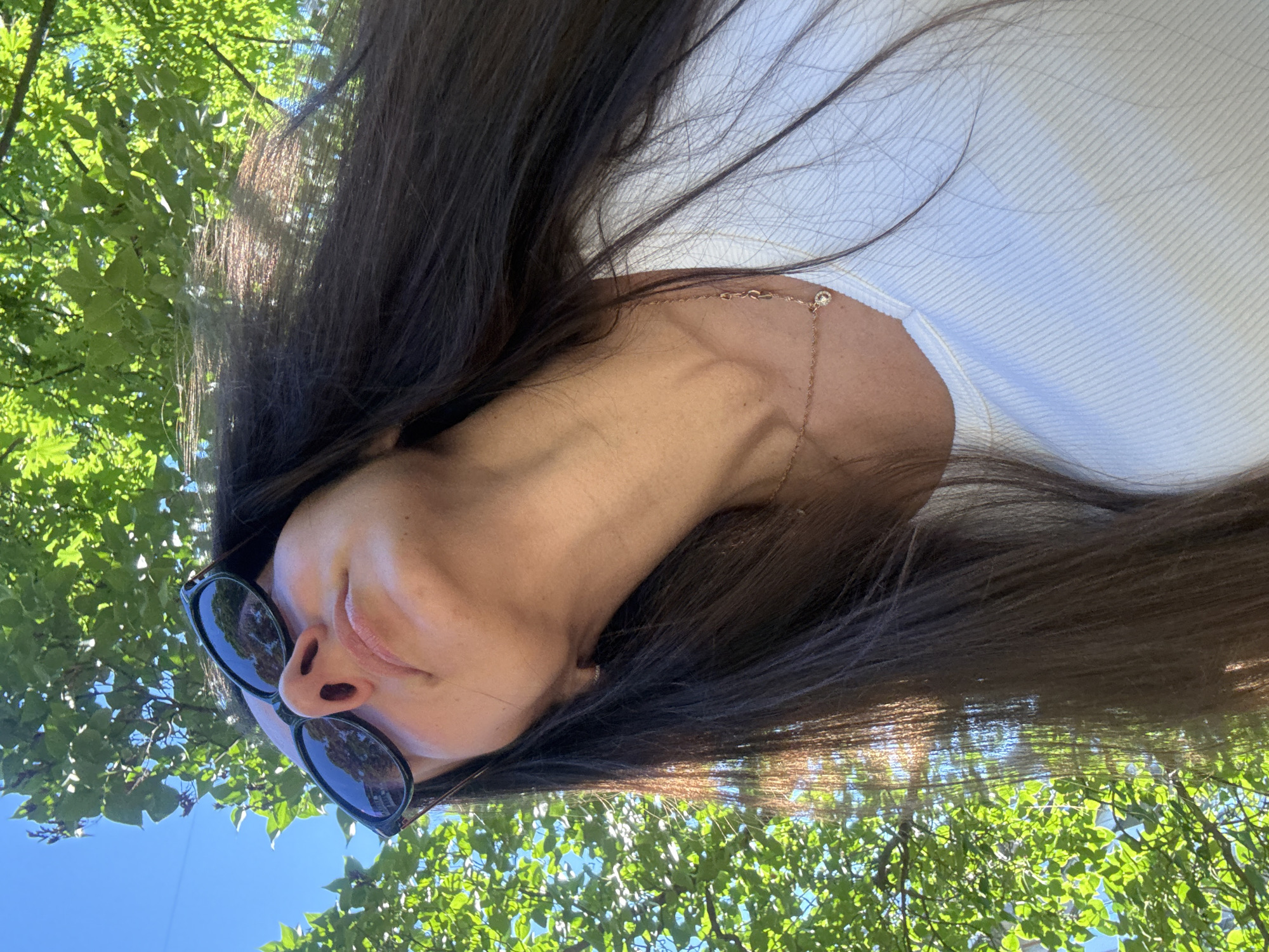 A close-up, low-angle shot captures a woman from the neck up, looking upwards towards a canopy of green leaves and a blue sky. She has long, dark brown hair that is partially illuminated by sunlight filtering through the trees. She is wearing sunglasses with dark lenses and a white, ribbed top. A delicate rose gold necklace with a small pendant rests on her collarbone. The scene appears to be outdoors, likely in a park or garden within Kyiv, Ukraine, given the lush greenery and bright sunlight suggesting a temperate climate. No specific landmarks or identifying features of Kyiv are visible, but the general environment is consistent with urban green spaces.
