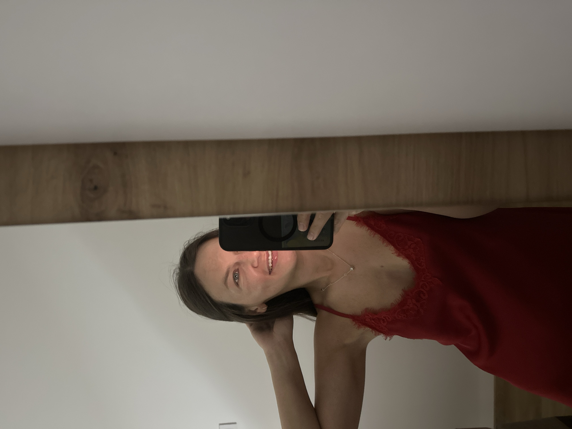 A woman is taking a selfie in a mirror. She is wearing a red slip with lace trim. She has her hair pulled back and is smiling. Her phone is visible in the mirror, and she is holding it to take the picture. The mirror is framed by a wooden border. The background is a plain white wall. The scene appears to be indoors, possibly in a bedroom or bathroom. The location in Donji Pobori, Montenegro is not discernible from the image.