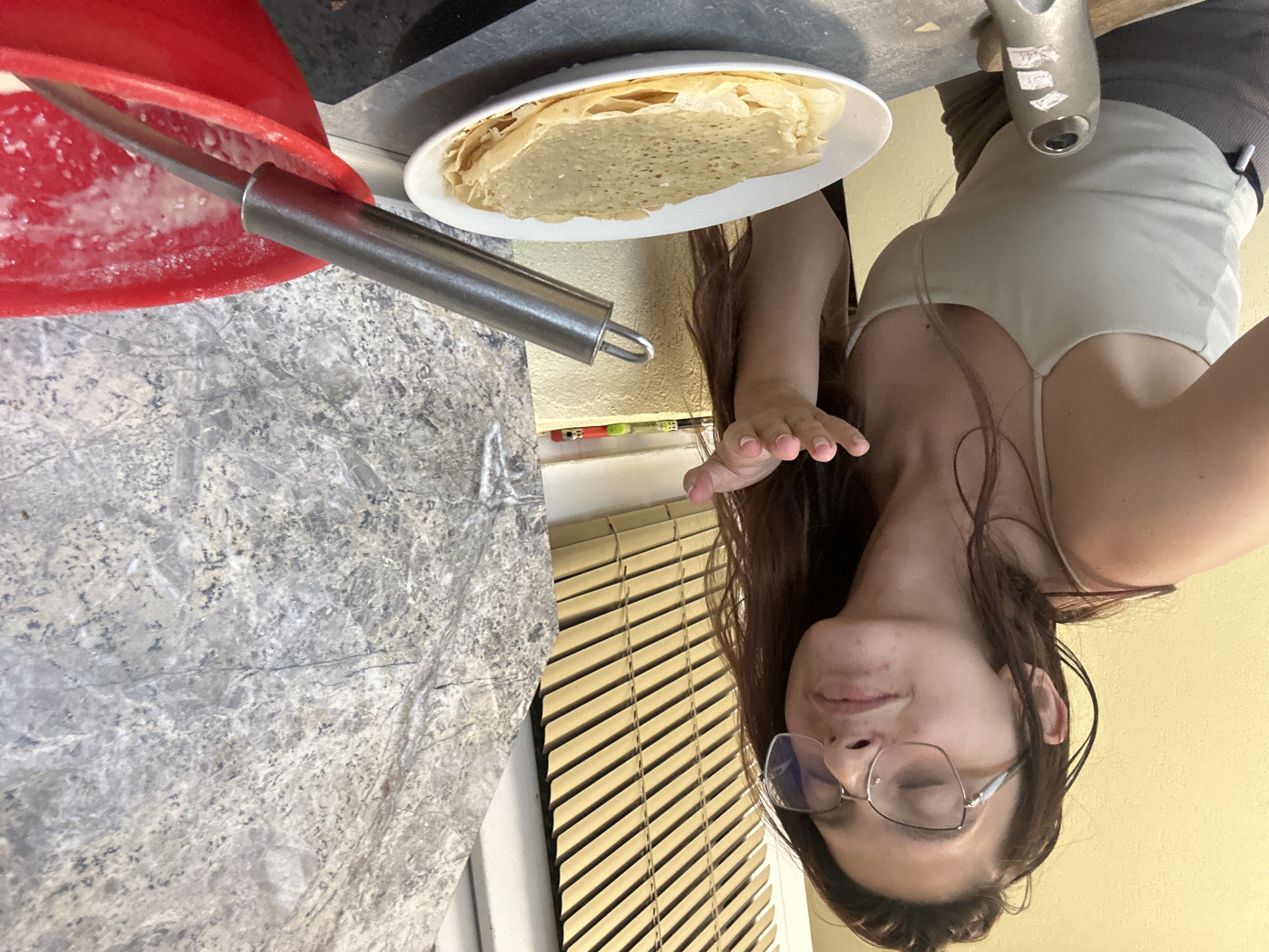 A young woman with long brown hair and glasses is in a kitchen, possibly in Tryavna, Bulgaria. She is wearing a light-colored tank top and darker shorts. Her eyes are closed, and she has a playful expression on her face as she blows a kiss with her right hand extended. In front of her, on a counter, is a white plate with a stack of thin, golden crepes or pancakes. To the right of the plate is a bright red bowl containing some white batter or liquid, with a metal whisk submerged in it. Behind the woman, there are blinds on a window and a textured, possibly granite, countertop or backsplash.