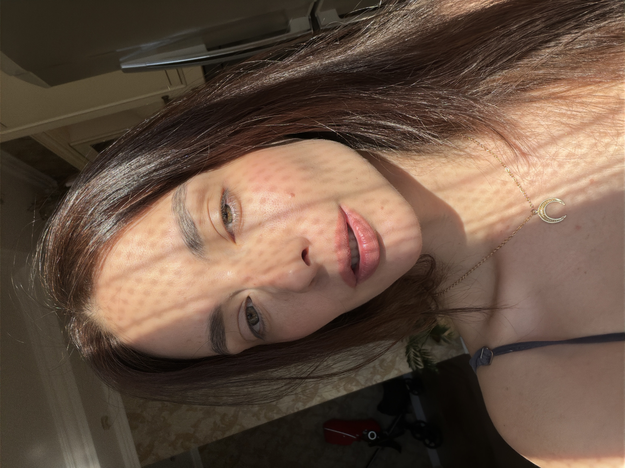 The image shows a close-up portrait of a woman with long, dark brown hair. She is looking slightly to her right, with her mouth slightly open. Her skin is fair, and she has a crescent moon necklace with small stones. The lighting suggests it is daytime, with sunlight casting a patterned shadow across her face. In the background, a stroller is visible in a dimly lit room with patterned wallpaper. This appears to be an indoor setting, possibly a home. There is no visible indication of the city of Ufa, Russia, or any specific urban environment.