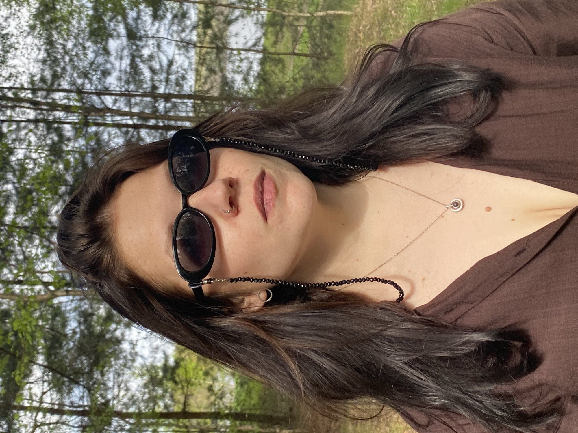 A woman wearing sunglasses and a brown top is outdoors with trees in the background. She is wearing a nose ring and a necklace. The location is not identifiable as Chaikovsky, Russia, based on the image.