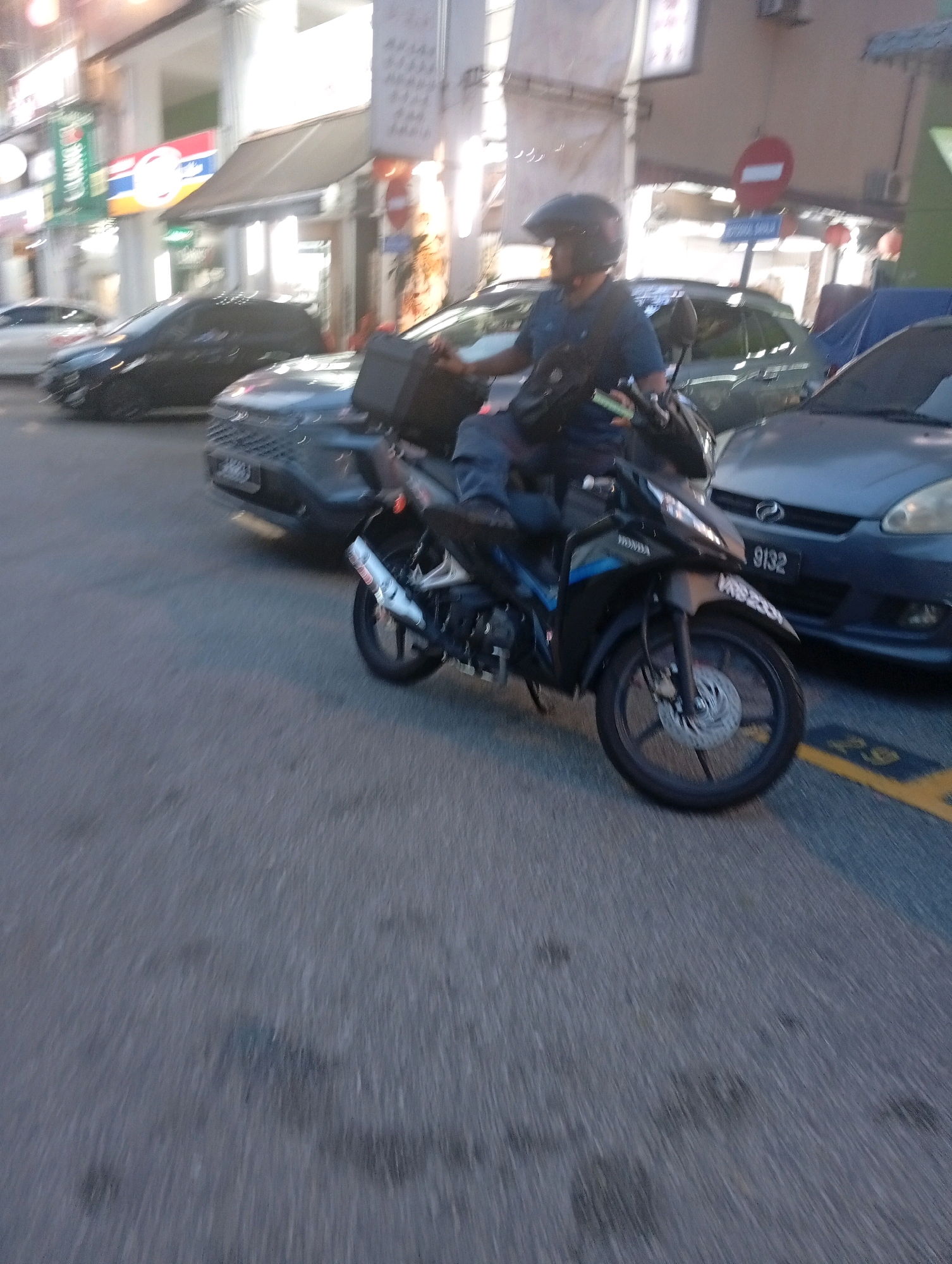 A motorcyclist wearing a black helmet and a dark blue shirt is riding a black and silver motorcycle on a paved road. The motorcycle has a black storage box attached to the rear. The rider is holding onto the handlebars with both hands and has a green object in their left hand. In the background, several cars are visible, including a dark-colored car in the foreground and a gray car with license plate "9132" behind the motorcycle. Buildings with awnings and signs are present on the left side of the image. The scene appears to be in an urban environment, likely a street in Selayang Municipal Council, Malaysia, given the presence of the specific car license plate format and general streetscape. Yellow road markings are visible on the asphalt. The image has a slight blur, indicating motion.
