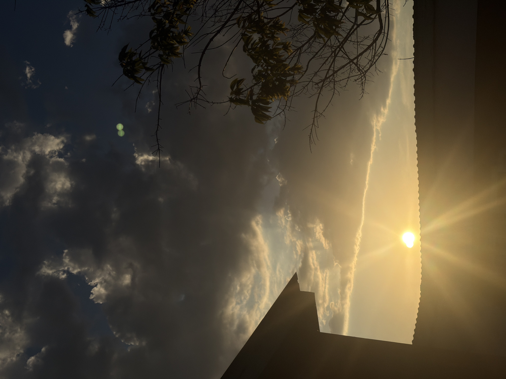 The image depicts a dramatic sky scene captured from a low angle, likely at dusk given the warm, golden light. The sun, a bright orb with visible rays, is positioned in the upper right quadrant, casting a warm glow across the sky. This light illuminates a strip of wispy clouds that stretch horizontally, contrasting with darker, more ominous clouds on the left side of the frame.
In the foreground, silhouetted against the bright sky, are the sharp angles of a building's roofline, suggesting an urban or suburban setting. To the left, branches of a leafless tree with sparse foliage hang into the frame from the top, adding a touch of nature to the scene.
A peculiar detail in the upper left quadrant is a pair of small, glowing green circles, resembling abstract shapes or perhaps a lens flare artifact. The overall mood is one of natural grandeur and perhaps a hint of foreboding due to the contrasting cloud formations.
There is no visible text in the image. The location context of Baurzhan Momyshuly, Kazakhstan, implies the scene is set in that region.