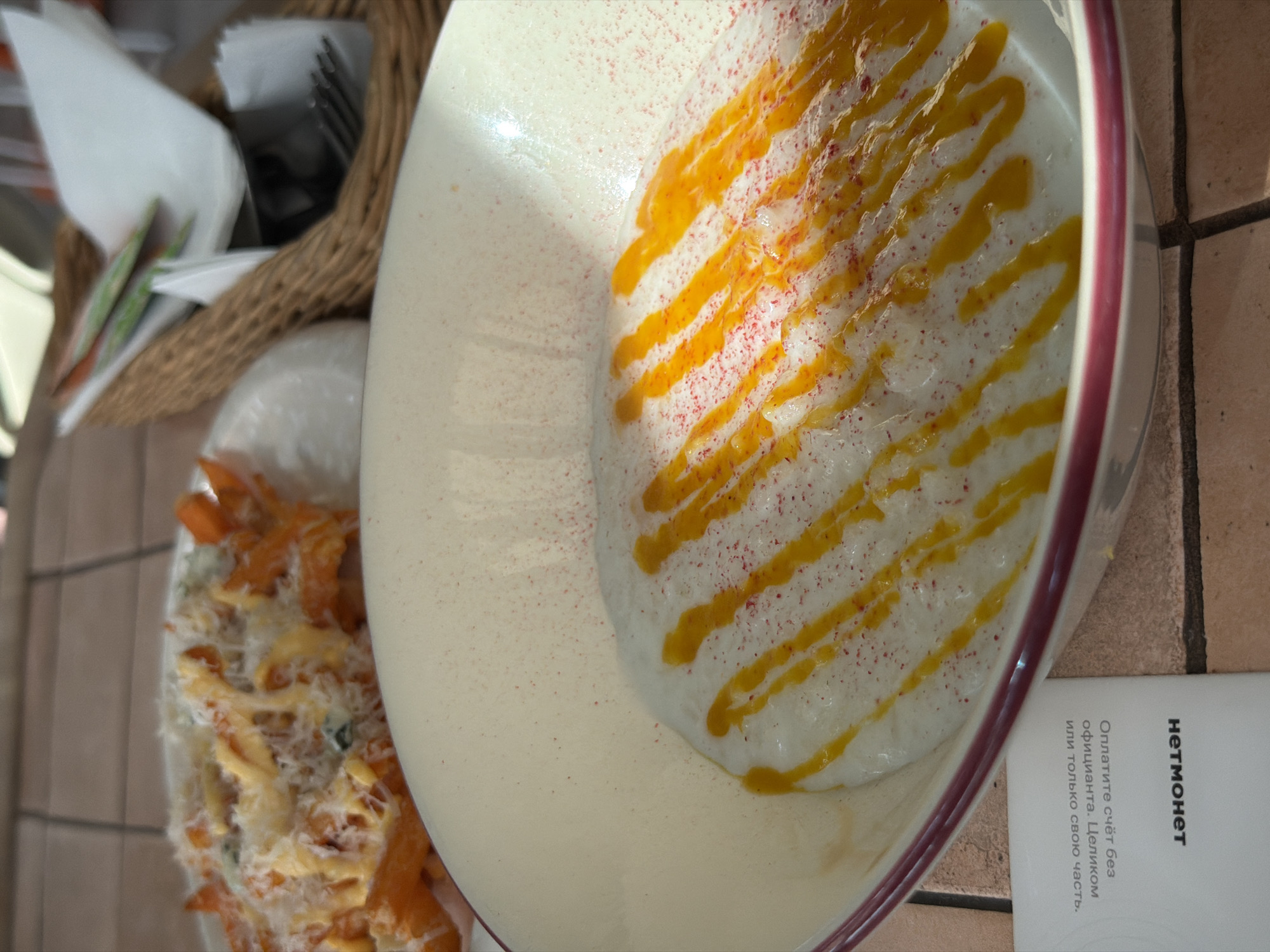 A close-up, slightly rotated overhead shot shows a bowl of creamy soup or porridge with lines of yellow sauce and a sprinkling of red specks. A side dish of cheesy, shredded food is visible next to the bowl. In the background, there's a woven basket holding cutlery and napkins. A card or receipt with the word "НЕТМОНЕТ" (NETMONET) and Russian text is partially visible on the right. The photo appears to be taken at a restaurant, as the scene is set on a table with a tiled surface. Considering the Russian text, it is likely the location is in Moscow, Russia.