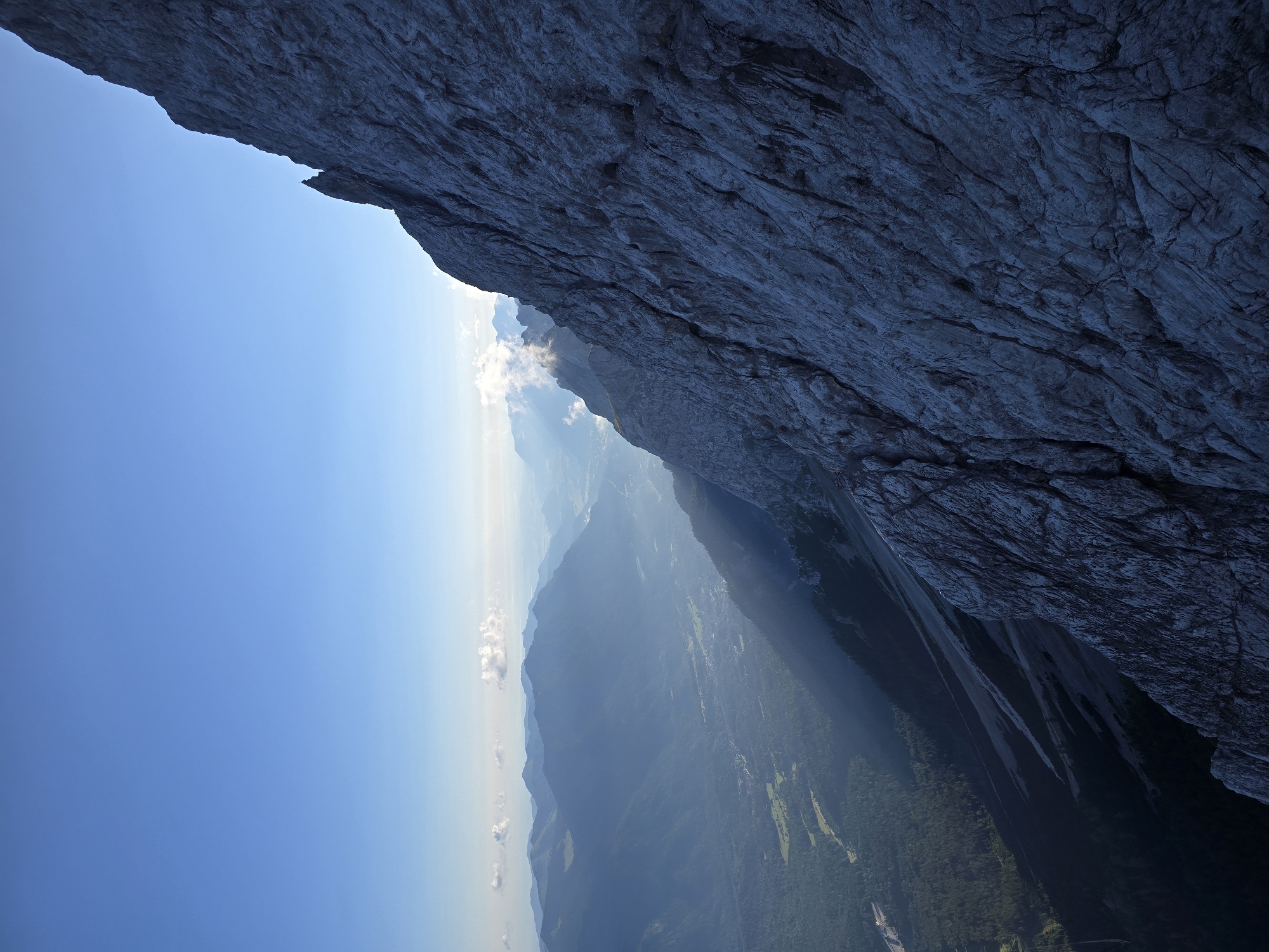 A dramatic, high-angle view captures a vast mountain landscape under a clear blue sky. The foreground is dominated by a sheer, rugged rock face, textured with deep fissures and shadowed crevices, suggesting a formidable mountain peak. This rock face occupies the right side of the frame, creating a strong diagonal line that leads the eye into the distance.
Below and to the left of the rock face, the scene opens up to a sprawling valley. Hazy mountains recede into the background, their forms softened by atmospheric perspective. Sunlight streaks through the atmosphere, illuminating parts of the valley floor and casting long shadows. The valley below appears to be a mix of dense green forests, cleared fields, and a small settlement with visible buildings. The overall impression is one of immense scale and natural grandeur.
This scene is likely from the Zugspitze massif, the highest peak in Germany, which is located near Grainau. The view is looking down from a significant elevation, offering a panoramic vista of the surrounding Bavarian Alps. There are no people or animals visible in the image. The environment is pristine and untouched, emphasizing the raw beauty of the alpine terrain.