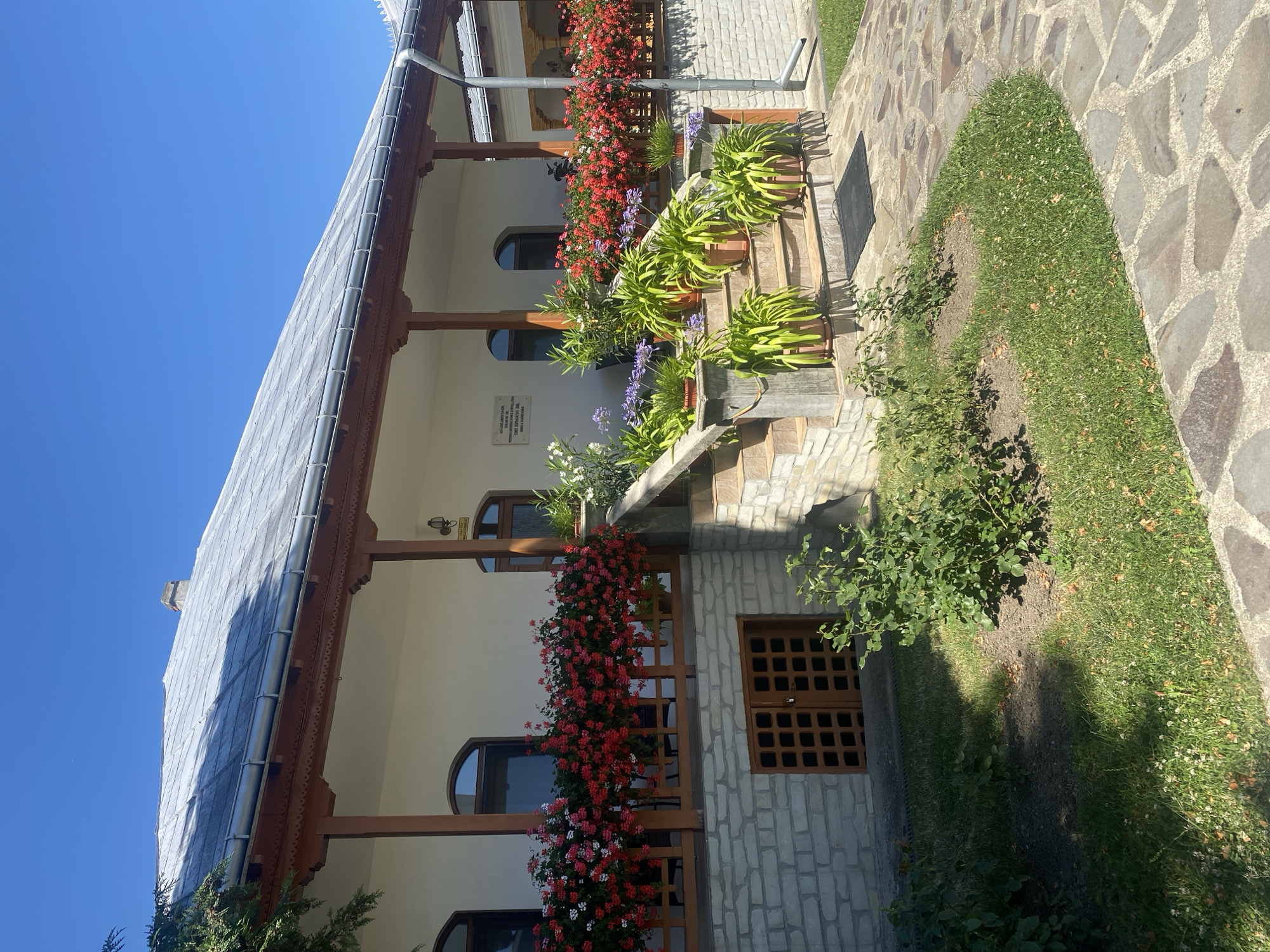 The image depicts the exterior of a building with a traditional Romanian architectural style, located in the village of Filioara, Romania. The building features a white facade, a grey tiled roof with wooden trim, and a covered porch supported by wooden columns. The porch is adorned with a railing filled with vibrant red and white flowers. A set of stone steps leads up to the porch, lined with potted plants, including large green leafy plants and purple flowers. The building's lower level is constructed with stone bricks and has a wooden door with a patterned design. The surrounding area is landscaped with grass, small bushes, and a stone pathway. The sky is clear and blue, indicating a sunny day. A sign is mounted on the wall of the building.