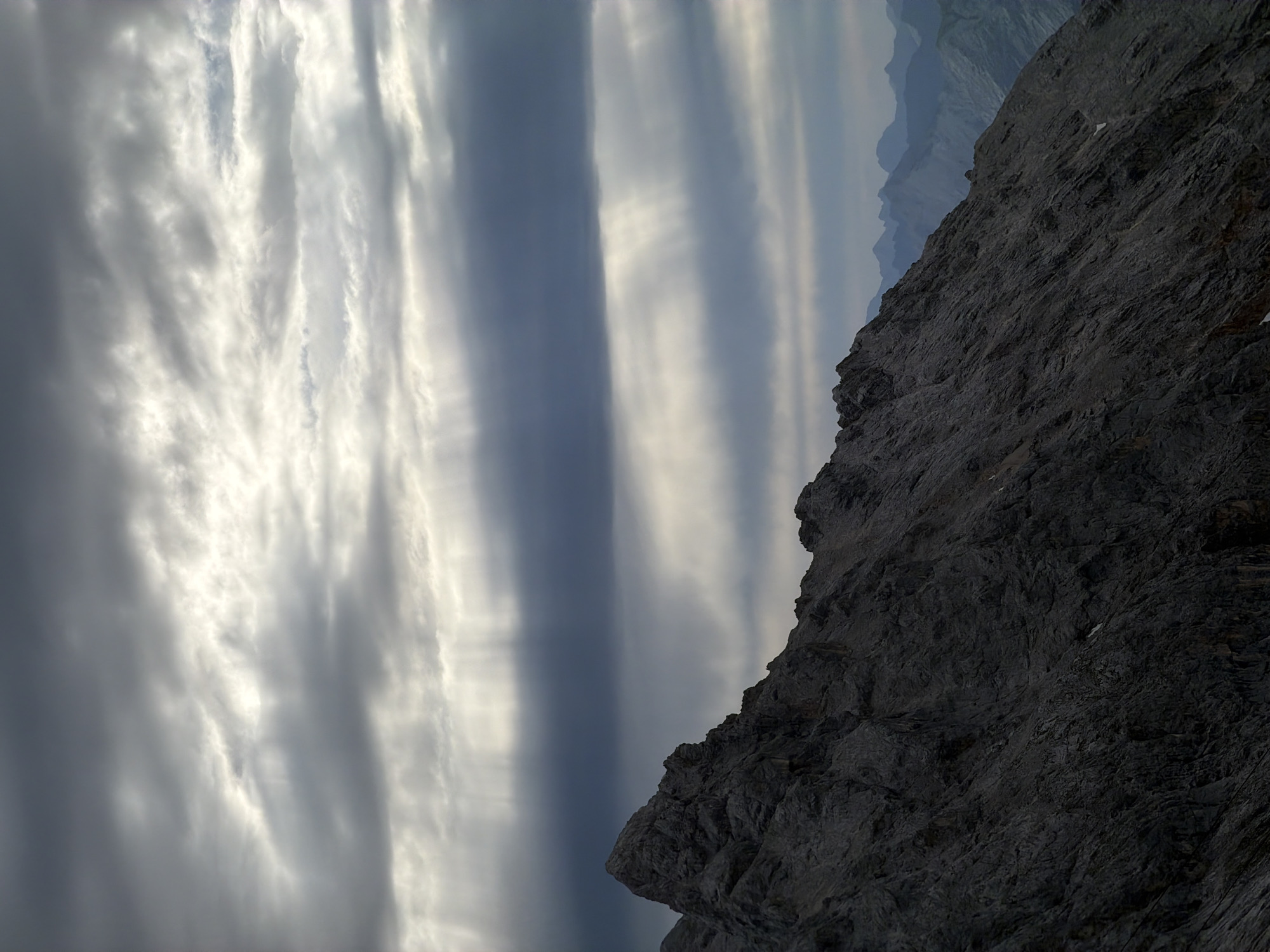 This is a photograph of a dramatic mountain landscape under a cloudy sky. The foreground is dominated by the rugged, dark grey rocky slope of a mountain peak, with sharp, jagged textures and visible strata. The slope descends from the left towards the right and into the distance. In the background, several layers of more distant, bluish mountain ranges are visible. The sky is filled with thick, textured clouds, with shafts of light piercing through in several places, creating a dramatic, almost ethereal effect. The overall lighting is subdued, suggesting either early morning, late evening, or overcast conditions. The image is a landscape shot, devoid of any human presence or activity. Given the jagged peaks and the scale, this scene is likely from the Zugspitze massif or a similar high-alpine region near Garmisch-Partenkirchen, Germany, known for its dramatic mountain scenery.
