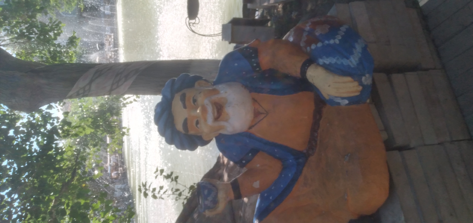 A statue of a smiling man wearing a blue robe and an orange tunic is positioned in front of a blurry fountain. The statue has a blue headpiece, a white beard, and a necklace. In his left hand, he holds a small blue object, possibly a cup. The fountain's water is illuminated by sunlight, creating a bright, sparkling effect. Trees with green leaves frame the scene on the left. The statue is leaning against a tree trunk that has a light-colored, patterned ribbon wrapped around it. The background also shows some wooden planks and what appears to be a metal structure. The overall impression is a bright, outdoor scene, likely during the daytime. No text is visible in the image.