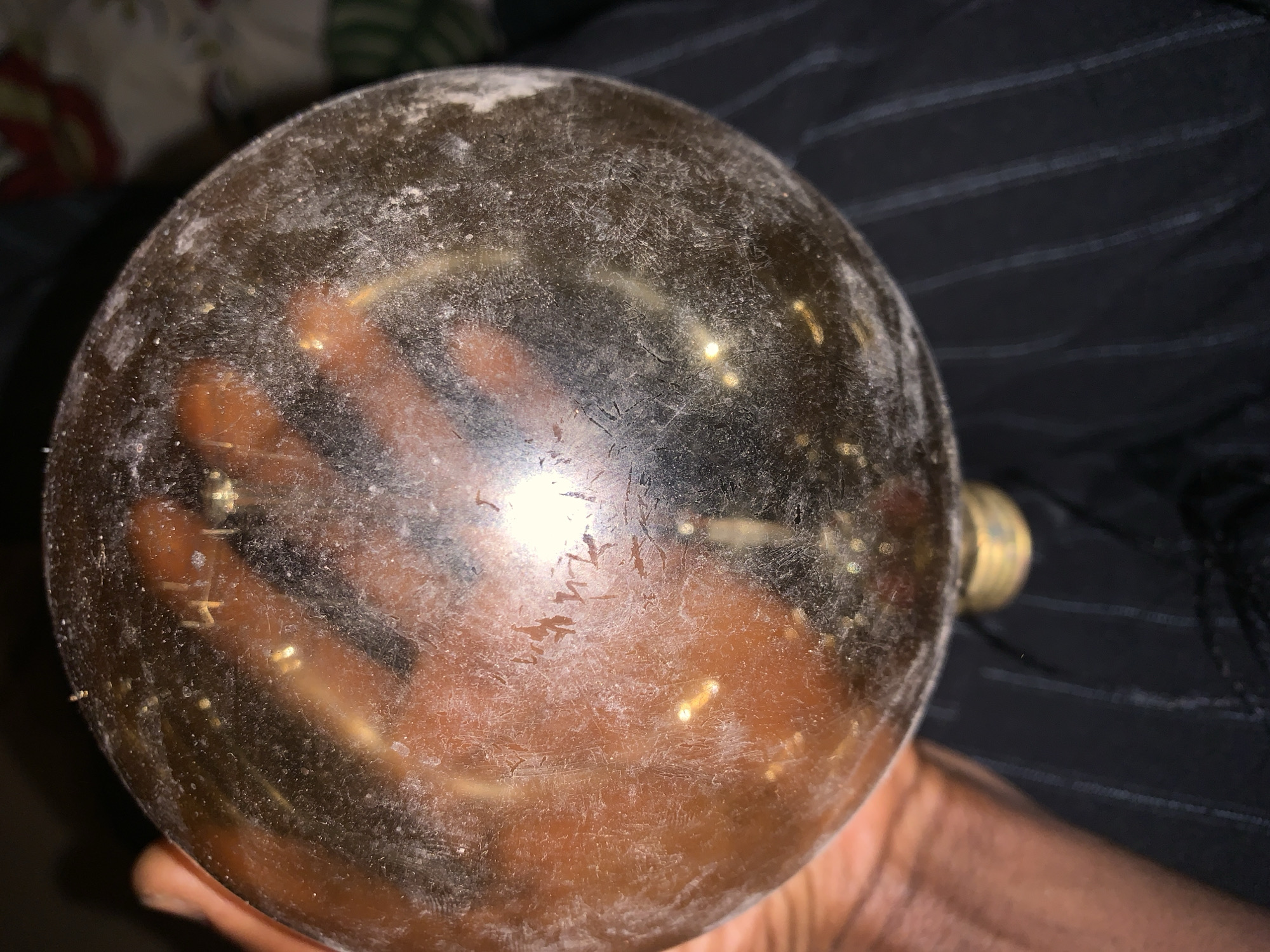 The image displays a large, spherical object, likely an old incandescent light bulb, held in a person's hand. The bulb is heavily discolored, appearing a murky dark brown or grey, and its surface is covered in visible scratches, smudges, and a layer of what appears to be dust or grime. Bright reflections, including a prominent central glare and several smaller glints, are visible on the bulb's curved surface, partially obscuring the interior.
A person's dark-skinned hand is holding the bulb from below, with the fingers visible through the translucent, discolored glass, appearing somewhat distorted. The brass screw base of the bulb is partially visible on the right side.
The background on the left is dark and out of focus, while the background on the right, behind the hand, shows a dark fabric with subtle, faint vertical stripes, possibly clothing or upholstery. The lighting conditions suggest an indoor environment with artificial light sources causing the strong reflections. Faint, angular scratched markings or symbols are visible on different areas of the bulb's surface, but no definitive, readable text can be discerned. This scene is located in Adankolo, Nigeria.