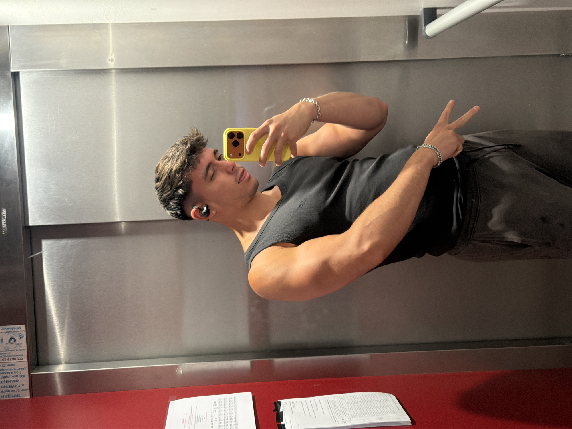 The image shows a young man taking a mirror selfie in what appears to be an elevator. He is wearing a black tank top and dark pants, with black earbuds in his ears and a silver bracelet on his wrist. He is holding a yellow smartphone to take the picture and is making a peace sign with his right hand. The elevator walls are metallic and reflective. There is a white bar or handle across the top of the elevator. On the bottom left of the image, there is a sticker with some text in what appears to be Cyrillic script, and some papers are visible on a red surface below the elevator doors. The lighting suggests it could be daytime. The man's expression is relaxed.