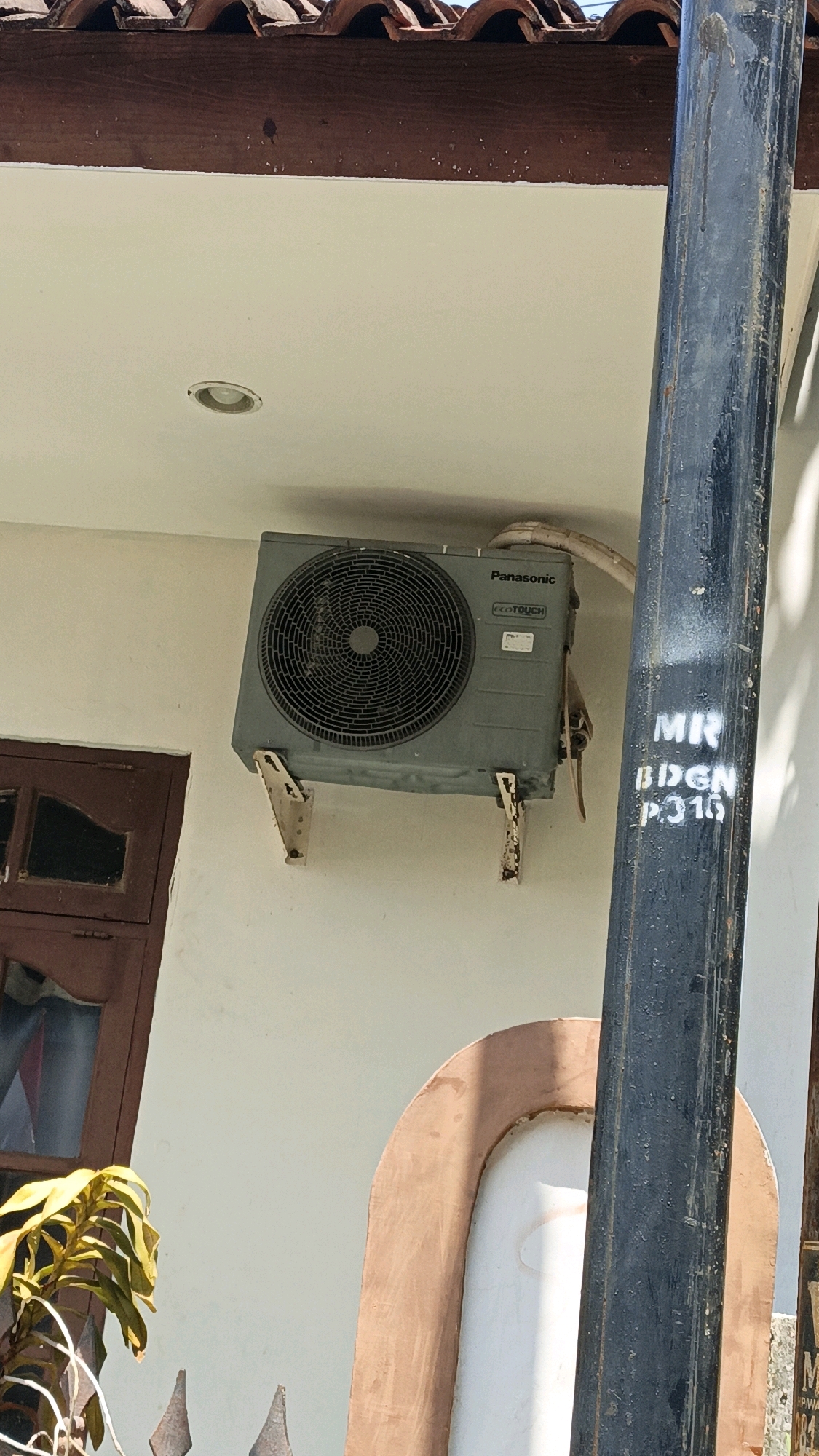 An outdoor image shows a gray Panasonic air conditioning unit mounted on a white wall with a dark brown tiled roof and wooden fascia above. The unit is supported by two rusty metal brackets. To the left of the air conditioner is a dark brown window with two panes. A black metal pole stands to the right of the air conditioner, with white spray-painted text reading "MR BDGN P.316" on it. In the lower right portion of the image, part of a white and brown arched structure is visible. A plant with green and yellow leaves is in the lower left corner, partially obscuring a decorative metal fence. The scene appears to be a residential building exterior in Cikal, Indonesia.