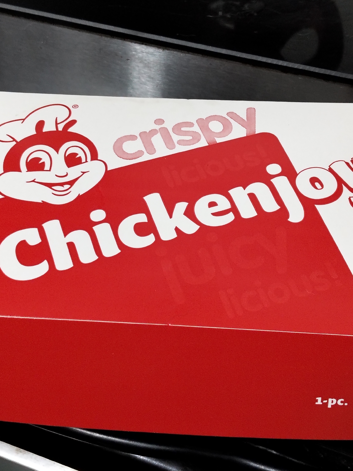 The image shows a close-up of a red Jollibee Chickenjoy box. The Jollibee mascot is visible on the left side of the box, wearing his chef's hat and smiling. The words "crispy" and "Chickenjoy" are prominently displayed in white lettering. The box appears to be new and unused. The background is dark and out of focus, suggesting the photo was taken indoors. The lower right corner of the box shows the text "1-pc." indicating the contents. The image does not provide any information about the specific location in Davao City, Philippines.