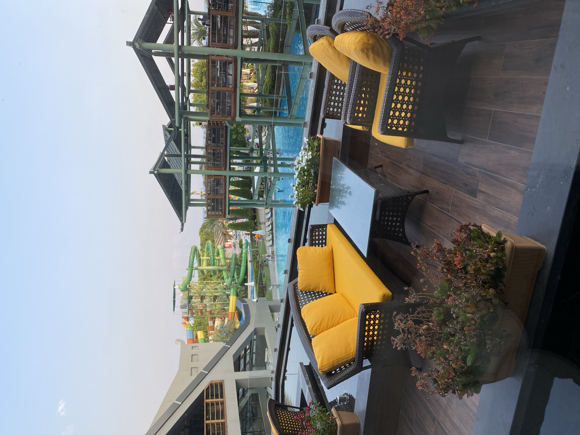 A high-angle outdoor shot shows a seating area on a tiled balcony overlooking a water park in Aksu, Turkey. The balcony features a dark wicker sofa with bright yellow cushions and three matching armchairs, all surrounding a glass-top coffee table. In front of the seating area, a planter holds a low-growing succulent-like plant with red flowers. To the left, another planter contains white flowers. Beyond the balcony, a large blue swimming pool is visible, with several people swimming and relaxing. Towering above the pool are colorful water slides, characteristic of a water park. In the background, a modern building with a distinctive angular roof is visible, and to the right, a multi-level structure with wooden accents and a green roof, likely part of the water park's facilities. The sky is a clear light blue, suggesting it is daytime.