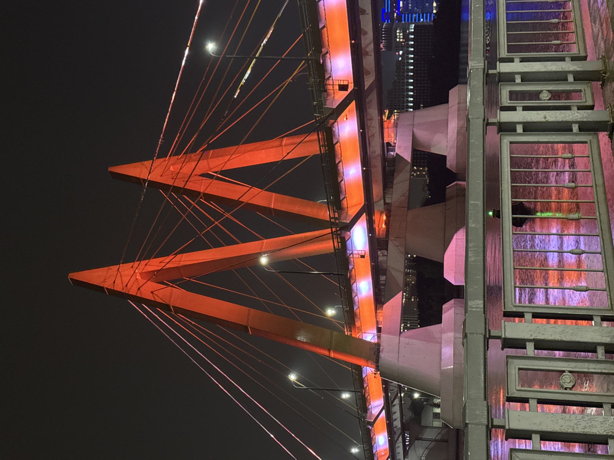 The image shows a cable-stayed bridge illuminated with orange and pink lights at night. The bridge has two prominent triangular pylons that are lit in orange. Numerous cables extend from these pylons, supporting the bridge deck, which is also illuminated with orange and pink lights. In the foreground, there is a metal railing along the waterfront, with reflections of the bridge lights on the water's surface. In the background, a city skyline with buildings and lights is visible. This is the Millennium Bridge in Kazan, Russia.