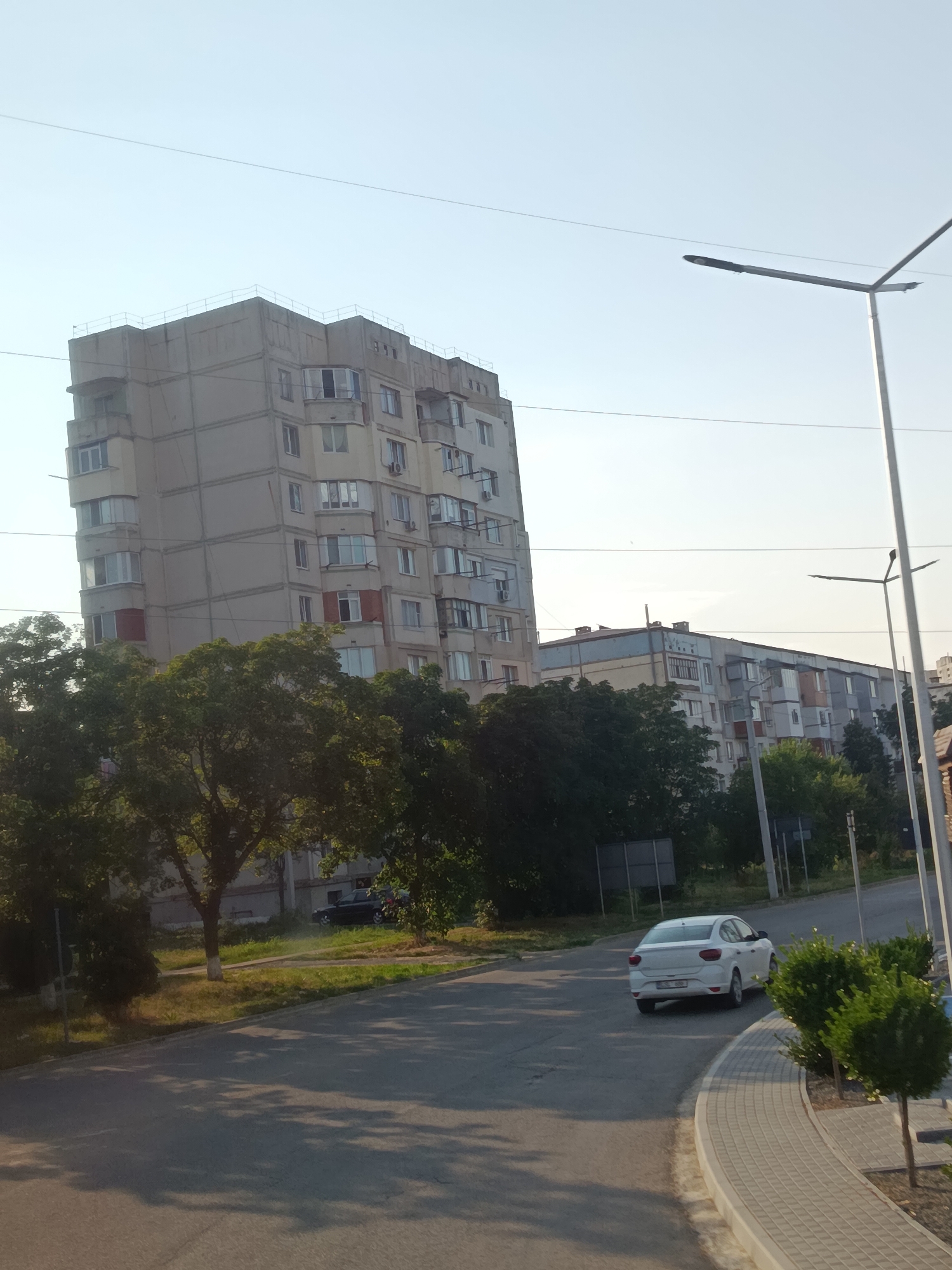 The image depicts a street scene in Cahul, Moldova, on a clear day with bright sunlight casting shadows. In the foreground, a gray asphalt road curves to the right, bordered by a light-colored paved walkway and green bushes. A white sedan is visible driving away from the viewer on the road. Behind the road, a line of lush green trees separates the street from a series of apartment buildings. The most prominent building is a tall, gray, multi-story structure with numerous windows and balconies. To the right of this building, a shorter, longer apartment building with a light blue and yellow facade is visible. Streetlights are present on poles along the roadside, and overhead wires crisscross the sky. A dark-colored car is parked in the distance near the trees, adjacent to one of the buildings. The overall environment suggests a residential area within a city.