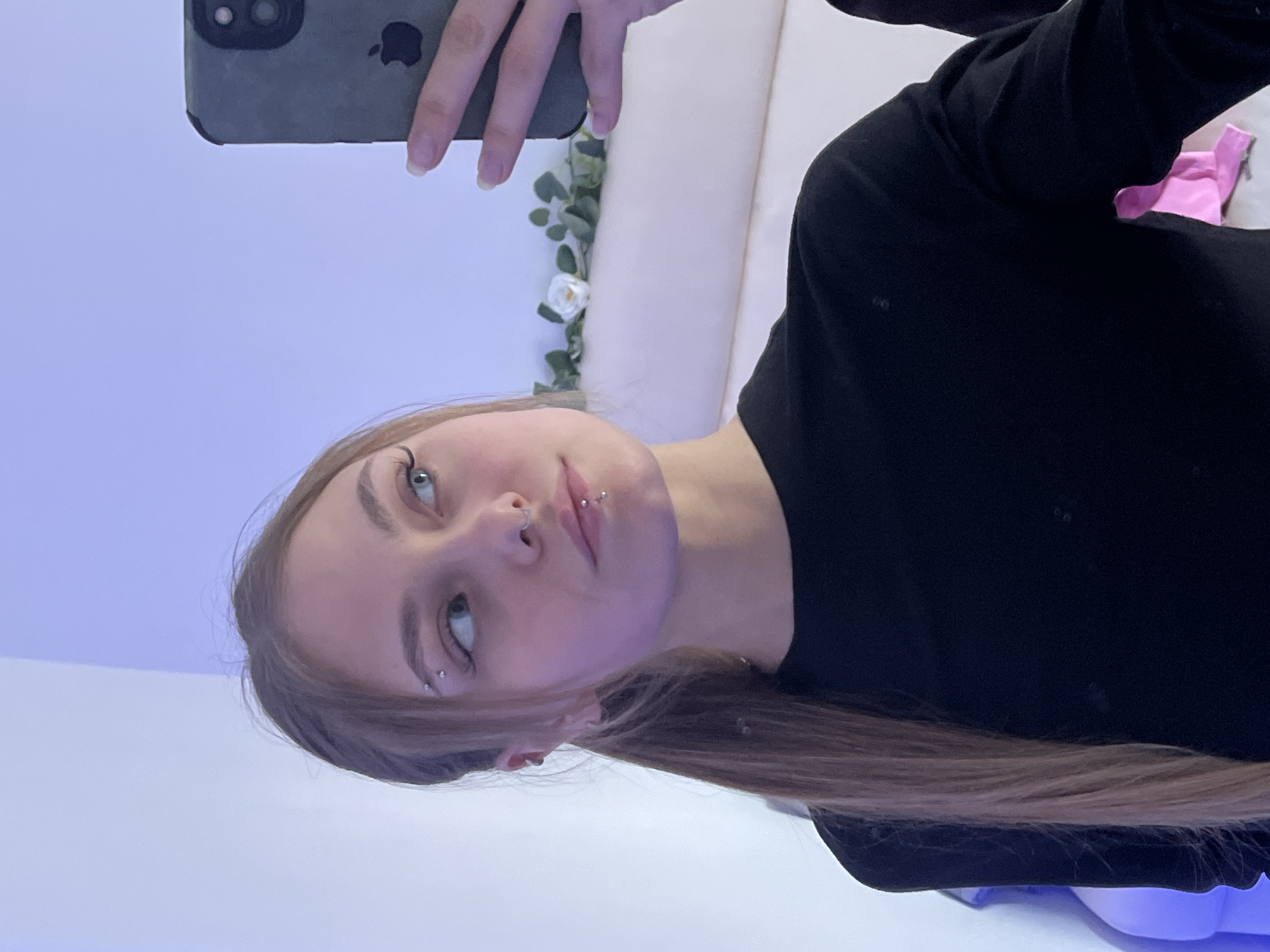 A young person with long, light brown hair, fair skin, and light-colored eyes is captured in a close-up selfie. The image is framed as if the person is looking upwards towards the camera, which they are holding with one hand, revealing an iPhone in a dark case with the distinct Apple logo visible on its back.
The person is wearing a plain black long-sleeved top. Notable facial features include several piercings: a septum piercing, a philtrum (medusa) piercing above the upper lip, and an eyebrow piercing. Their expression is neutral, with a slight upward gaze.
The background suggests an indoor setting, likely a room. A light-colored wall or furniture, possibly a headboard, is visible behind them. Adorning the wall is a decorative element consisting of green leafy garland intertwined with small white artificial flowers. A subtle blue or purple artificial light source casts a hue on the background, contributing to the ambiance. A small, crumpled pink object is partially visible in the upper right corner of the background, hinting at other items in the room. This scene is contextualized as taking place in Фоки, Russia.