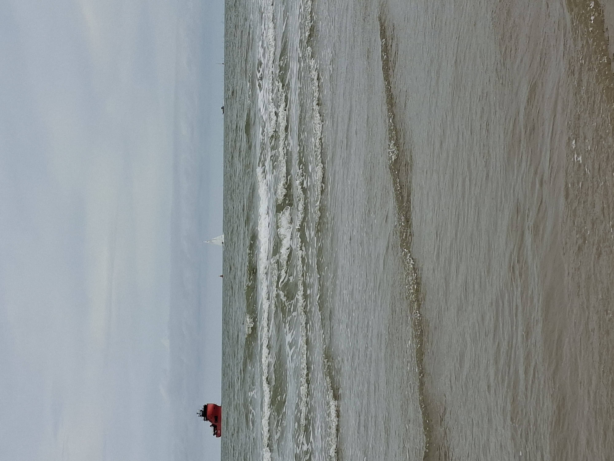 The image depicts a wide, calm sea under a cloudy sky. In the distance, a large red ship is visible on the left side of the frame, while a white sailboat is positioned to the right of the red ship. Further to the right, a small dark boat is also seen on the water. The foreground shows gentle waves lapping onto a sandy beach, suggesting a coastal environment. The overall atmosphere is serene and somewhat overcast. This scene is consistent with the North Sea coast of Ostend, Belgium, a popular seaside resort city known for its long beaches and maritime activity.