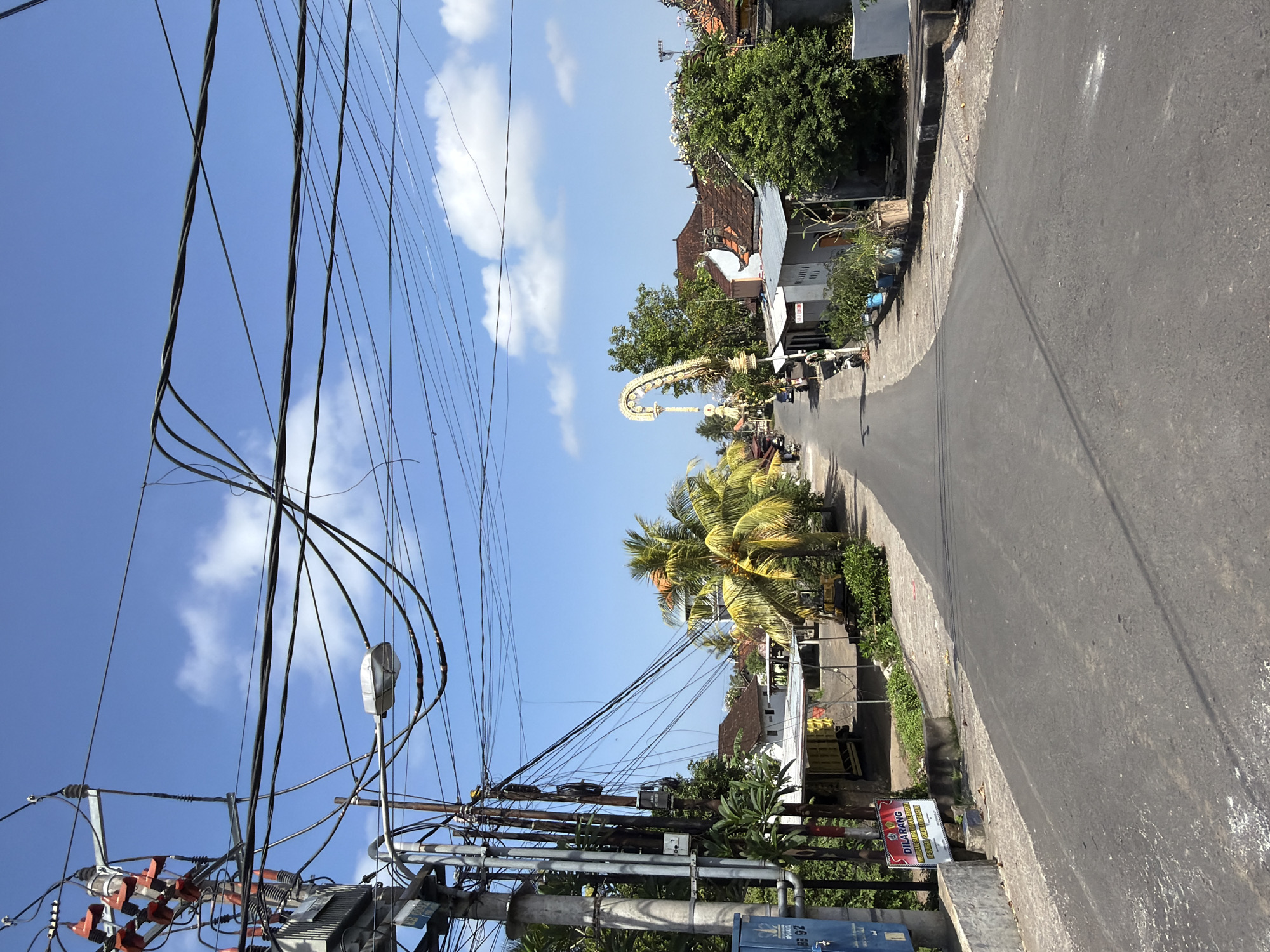 The image shows a street scene in Blahbatuh, Indonesia, under a clear blue sky with scattered clouds. A paved road extends into the distance, lined on both sides by buildings and vegetation. On the left side of the road, there is a prominent utility pole with a complex network of electrical wires and a transformer. A blue electrical box is attached to the base of the pole, and a red and white sign reading "DILARANG MEMASANG IKAN DI TEMPAT UMUM" is posted nearby. Further down the street, palm trees with yellowing fronds are visible. On the right side of the road, houses with tiled roofs and lush green trees are present. In the distance, an ornate archway with decorative elements is visible, suggesting a cultural or ceremonial marker. A few parked motorcycles and a yellow excavator are also present in the scene. The overall impression is a typical street in a tropical, possibly developing, urban or suburban area.