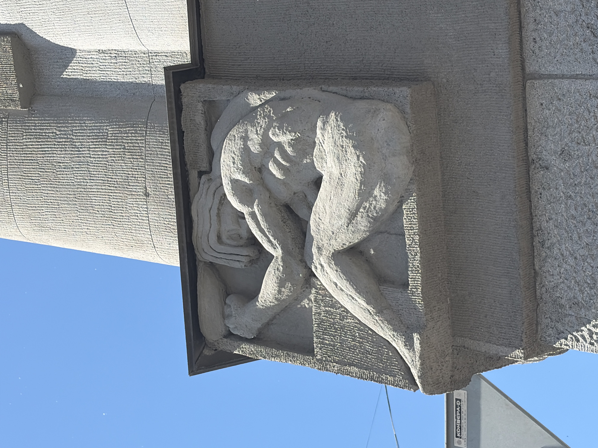 This media file features a stone relief sculpture of a person in a distressed or contemplative pose. The sculpture is attached to a building made of textured stone, with some areas showing a rougher granite finish. The person in the relief is hunched over, with their arms wrapped around their head, suggesting a state of sorrow, pain, or deep thought. The carving is detailed, showing musculature and facial features. The sculpture is mounted against a bright blue sky, indicating a clear day, likely during daylight hours. The angle of the shot suggests the viewer is looking up at the sculpture. Some structural elements of the building are visible, including rough-hewn stone blocks and a darker, possibly metal, frame around the relief. A faint shadow is cast by an overhang onto the building's facade. Partially visible at the bottom right is a grey metal structure with some text on it. The text reads "KOHBEPA-D". Given the location context of Saint Petersburg, Russia, this sculpture is likely an architectural detail on a public building.