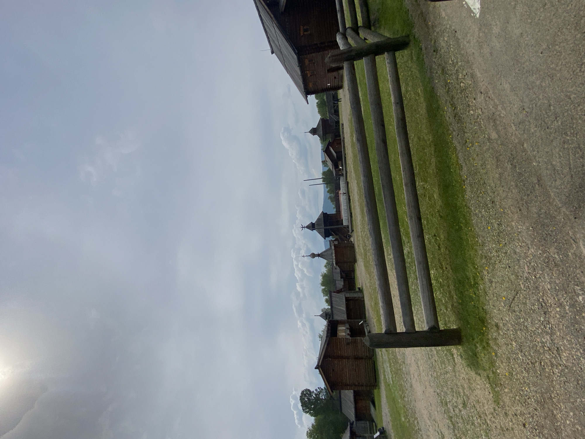 The image depicts an outdoor scene in the Taltsy (Irkutsk) museum-reserve in Russia, likely during a cloudy day. In the foreground, a weathered wooden fence with three horizontal logs stretches across the frame. Beyond the fence, a dirt and gravel path gives way to patches of green grass. Several traditional wooden structures, characteristic of Siberian architecture, are visible in the mid-ground and background. These include log houses with dark wooden walls and pitched roofs, some featuring visible windows and balconies. Distinctive domed structures with spires, possibly churches or watchtowers, punctuate the landscape. In the distance, a line of trees and rolling hills can be seen under a bright, overcast sky with some cumulus clouds. A few indistinct figures, presumably visitors, are scattered throughout the site.