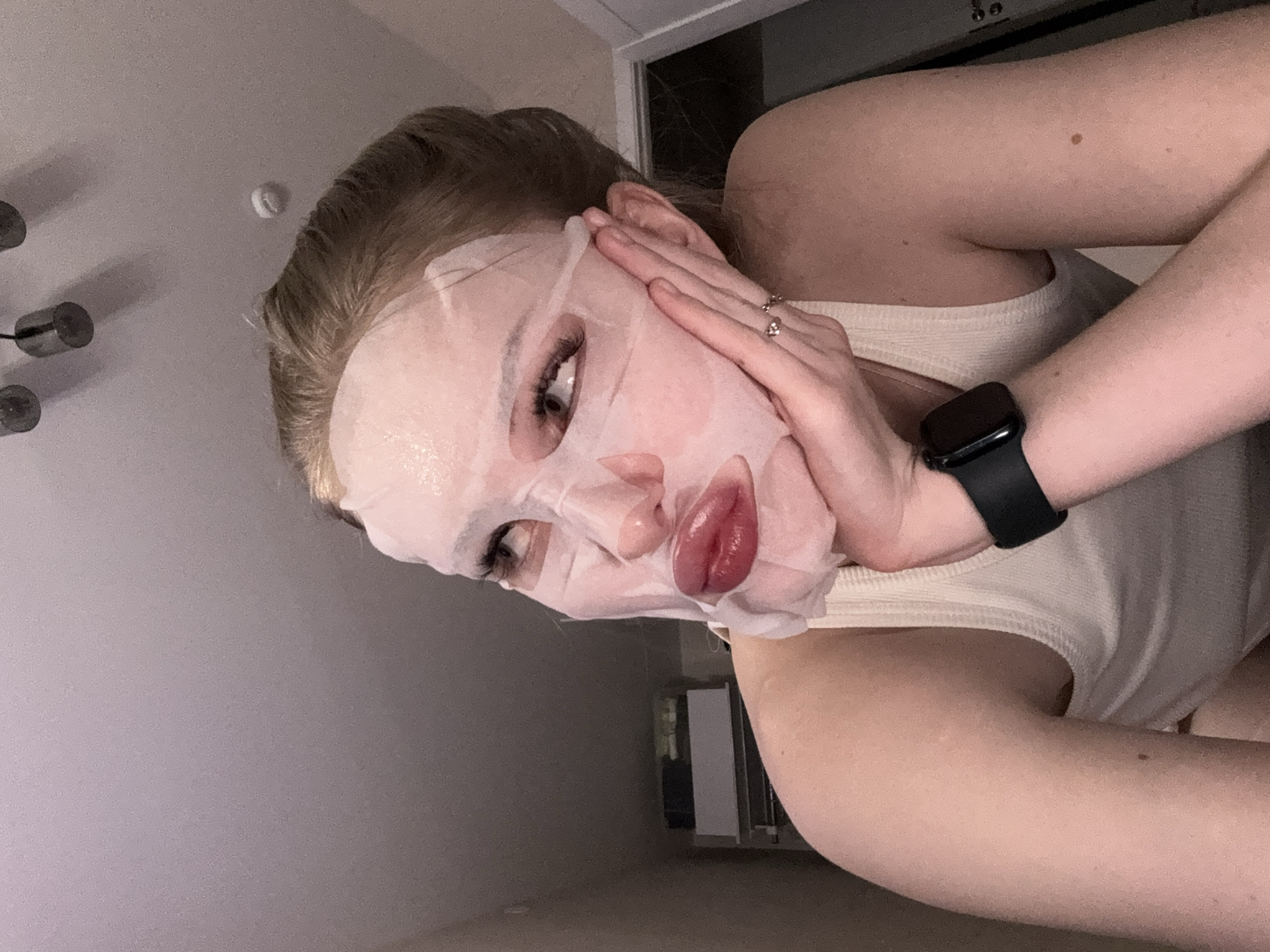 A young woman is depicted indoors, likely engaging in a skincare routine. Her face is mostly covered by a white sheet mask, which has cutouts for her eyes, nostrils, and mouth. Her eyes are visible through the mask, featuring dark, prominent eyelashes. Her lips, which appear full and glossy with a reddish tint, are also visible through the mask's opening.
She has light-colored hair, possibly blonde or light brown, which appears pulled back from her face. Her hands are positioned near her cheeks and ears, possibly adjusting the mask or posing. On her right hand, she wears several rings. A black smartwatch is visible on her left wrist. She is wearing a light-colored, ribbed tank top or camisole.
The setting appears to be a room within a home, with plain walls and a ceiling visible in the background. The lighting is dim, suggesting it could be evening or night. A part of a light fixture with cylindrical elements is discernible on the ceiling. There is no visible text in the image. The image is taken from a slightly low angle, looking upwards. While the provided context is Perm, Russia, there are no specific visual cues within the image itself to indicate this location.
