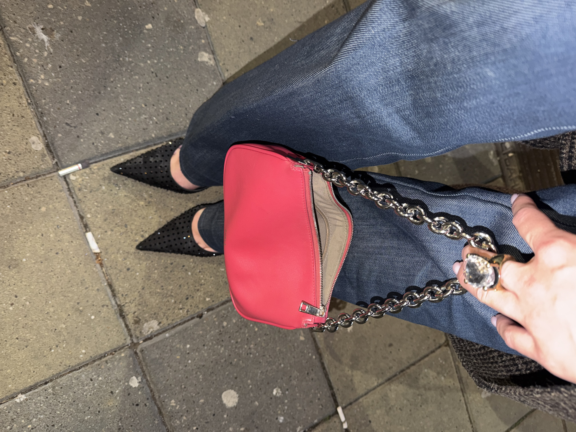 A top-down shot shows a person seated on a Belgrade, Serbia sidewalk. They are wearing dark-wash denim jeans and black, studded pointed-toe heels. A bright pink handbag with a silver chain strap rests on the person's lap. The bag is partially unzipped, revealing a light-colored interior. The person's hand, adorned with a large, square-cut ring, is visible on the right side of the frame. Two discarded cigarette butts lay on the pavement near the shoes. The sidewalk consists of gray, speckled tiles. The image is lit as though taken at night.