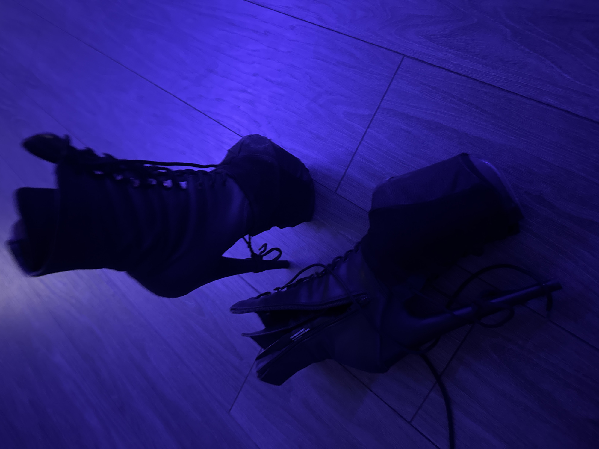 The image captures two dark-colored, high-heeled boots resting on a wooden floor, bathed in a strong, artificial deep blue or purple light. The lighting creates a distinct, moody atmosphere, suggesting an indoor setting perhaps at night or in a room with specialized illumination rather than natural daylight.
The boots appear to be a high-heeled, lace-up style, possibly resembling combat or fashion boots. The boot on the left clearly displays a tall, slender heel. The boot on the right, which is closer to the viewer and partially unzipped, also features laces and a prominent zipper compartment or pocket on its side. On this compartment, a small, white label or logo is faintly visible, though the text on it is indecipherable due to the low light and resolution. The wooden floor beneath the boots shows visible planks and grain patterns.
There are no people present in the image, and no specific activity is taking place; the boots are simply static objects on the floor. The scene conveys a sense of stillness and a stylized aesthetic due to the intense monochromatic lighting. No identifiable location cues other than the wooden floor are present.
